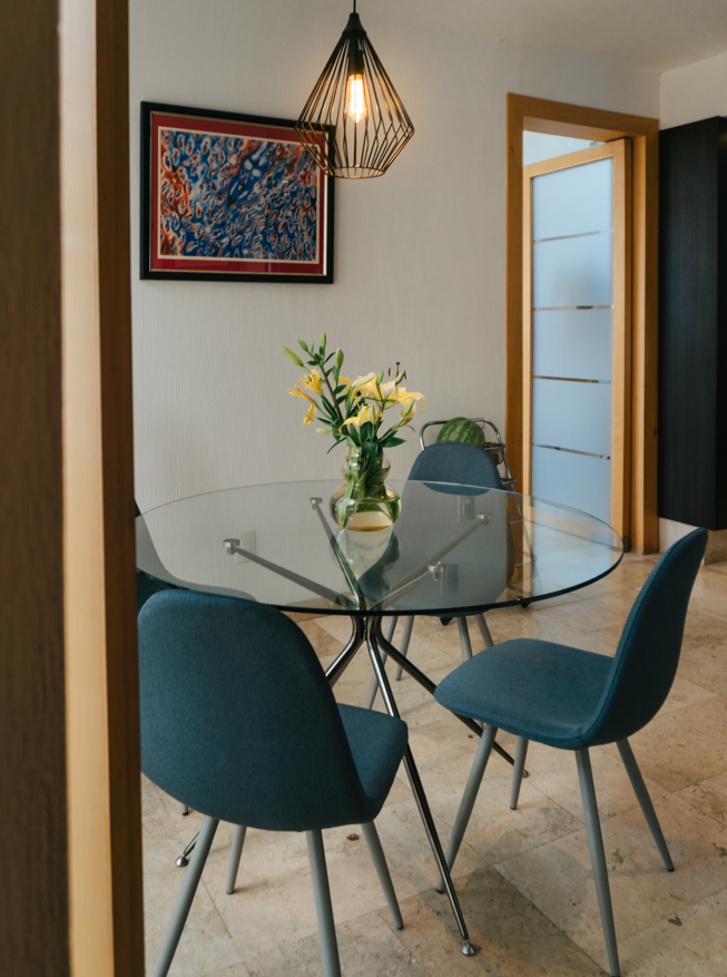Comedor moderno con mesa de vidrio y sillas azules, florero con flores amarillas, luz colgante y pintura colorida en la pared.