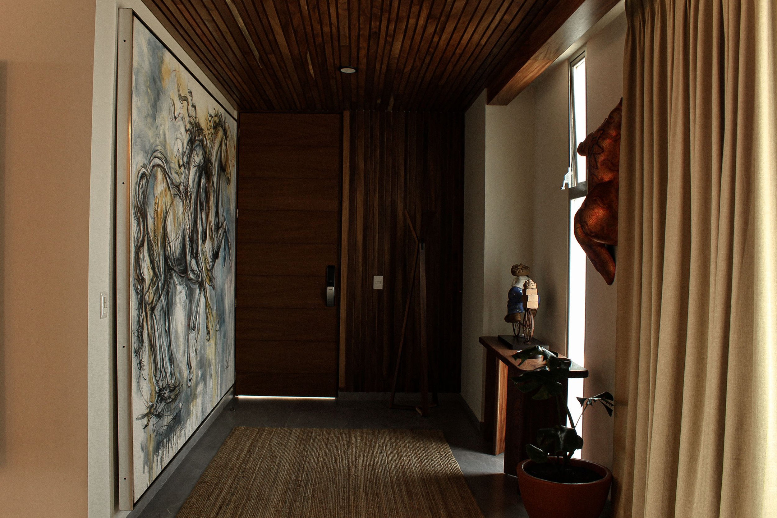 Vestíbulo interior con puerta de madera, pintura de caballos, tapete marrón, mesa de madera con figura decorativa y plantas en macetas.