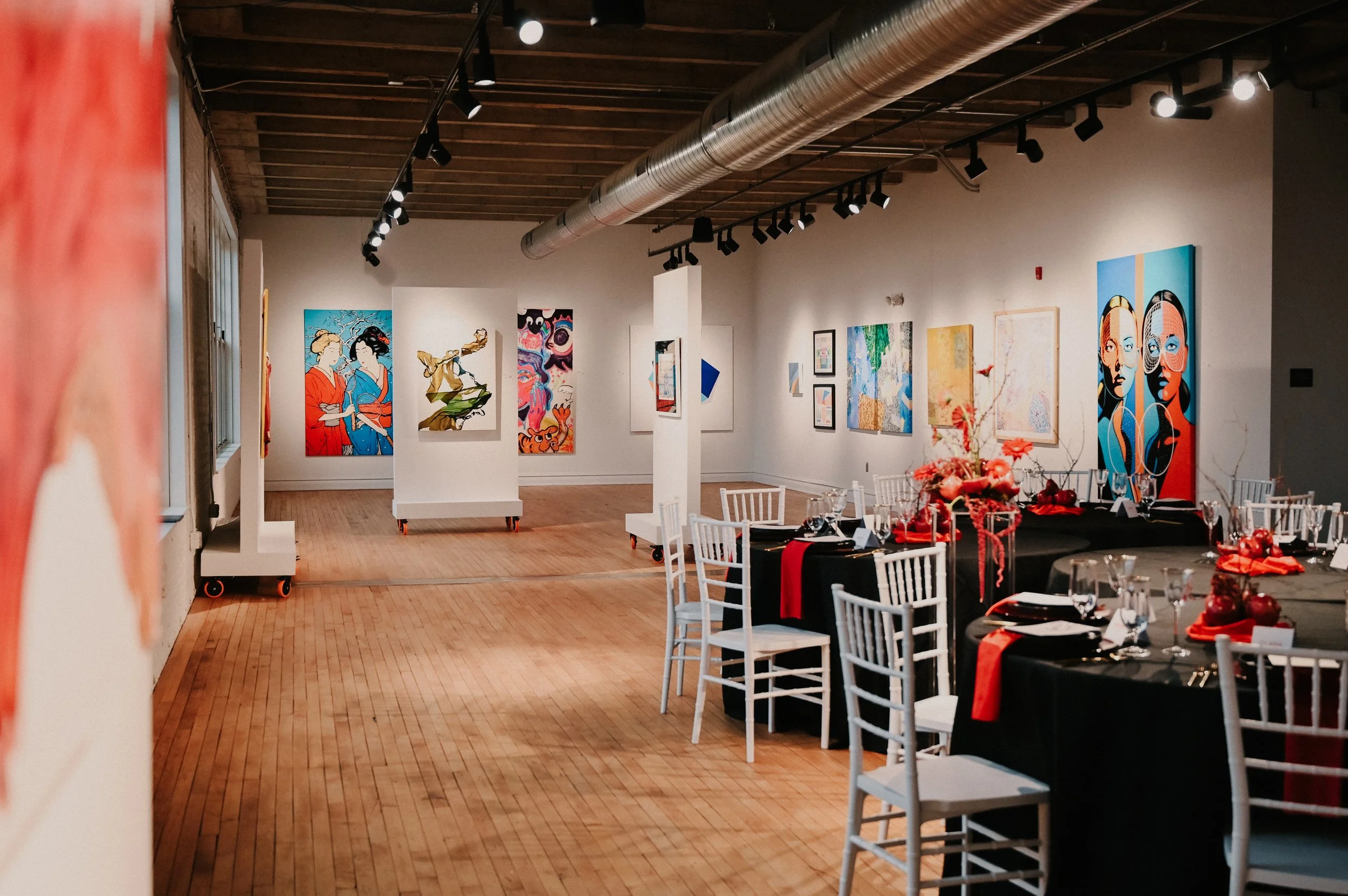 A contemporary art gallery with exposed wood floors and ceiling beams, featuring colorful artworks displayed on white walls and movable panels, alongside an elegant dining setup with white chairs, black table linens, red accents, floral centerpieces
