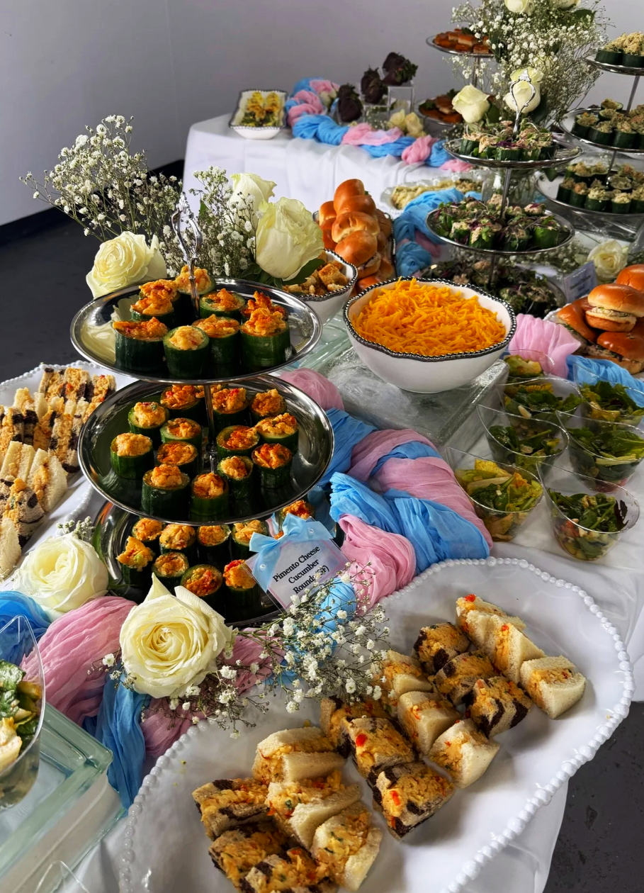 A table filled with various appetizers and snacks, including sushi rolls, cheese and cucumber bites, shredded cheese, baked goods, and salads, decorated with white flowers and pink and blue fabric drapes.