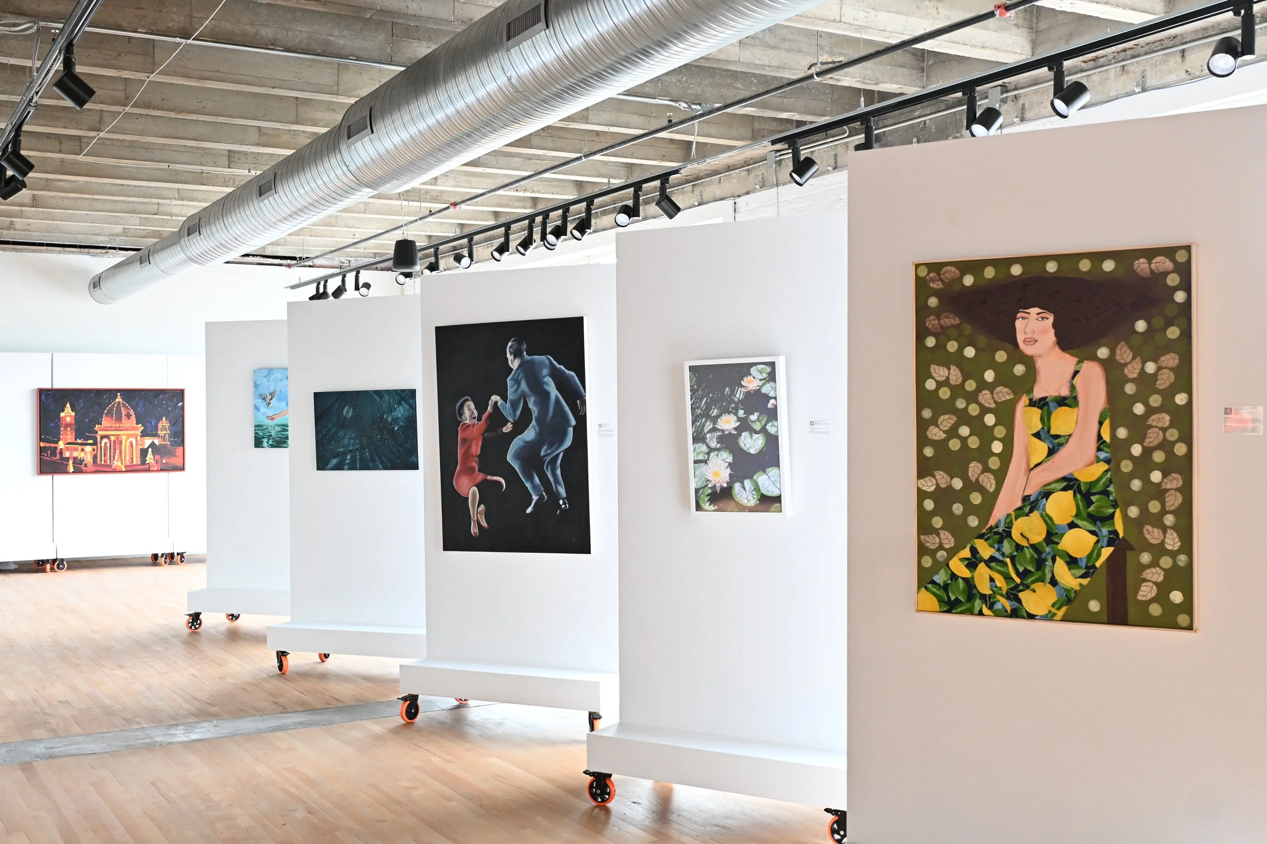 An art gallery with paintings displayed on white walls, including a woman with large hair and a colorful dress, a couple dancing, and various nature scenes. The gallery has a wooden floor, an industrial ceiling with exposed pipes and track lighting, 