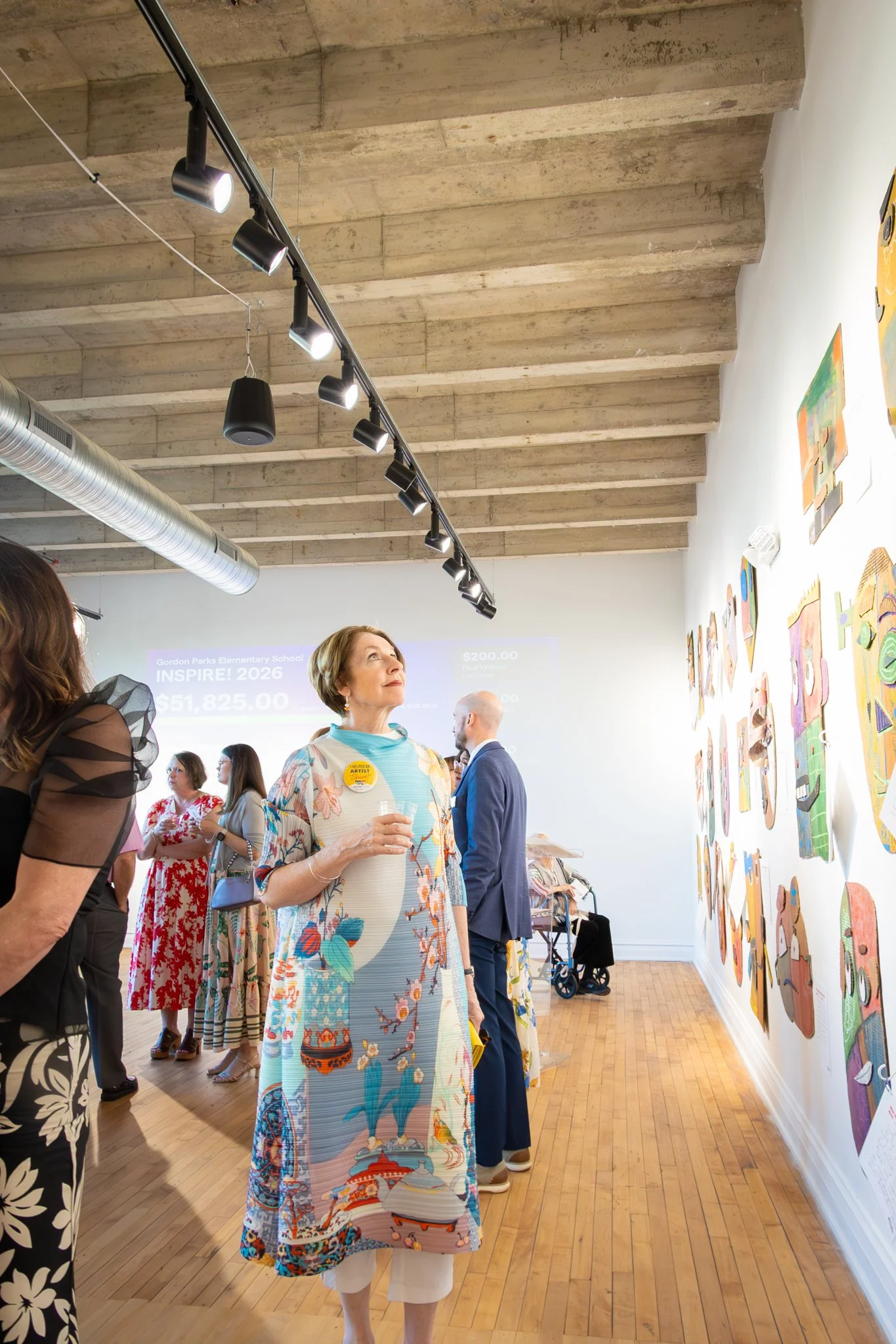 People attending an art exhibit in a gallery with wall-mounted abstract paintings, illuminated by track lighting, and a woman in the foreground wearing a colorful dress with floral and scenic patterns.