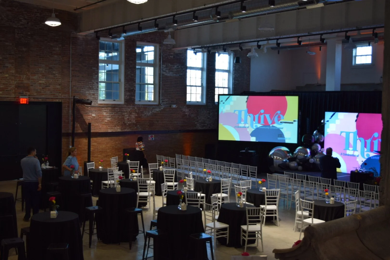 Event venue with tables and chairs, large screens displaying the word 'Thrive,' balloons, and a few people preparing for an event in a brick-walled space with high windows.