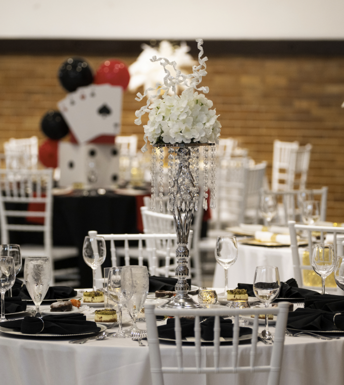 Elegant banquet table setting with a tall silver centerpiece, white flowers, and hanging crystal decorations, in a decorated event space.