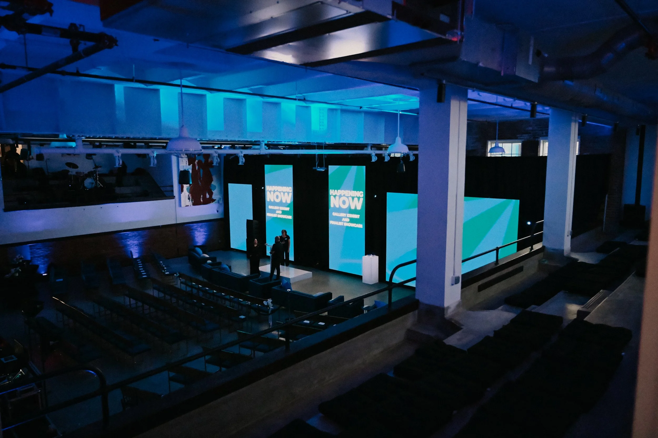 Indoor event space with large screens displaying 'HAPPENING NOW' and people standing on a stage, illuminated with blue lighting.
