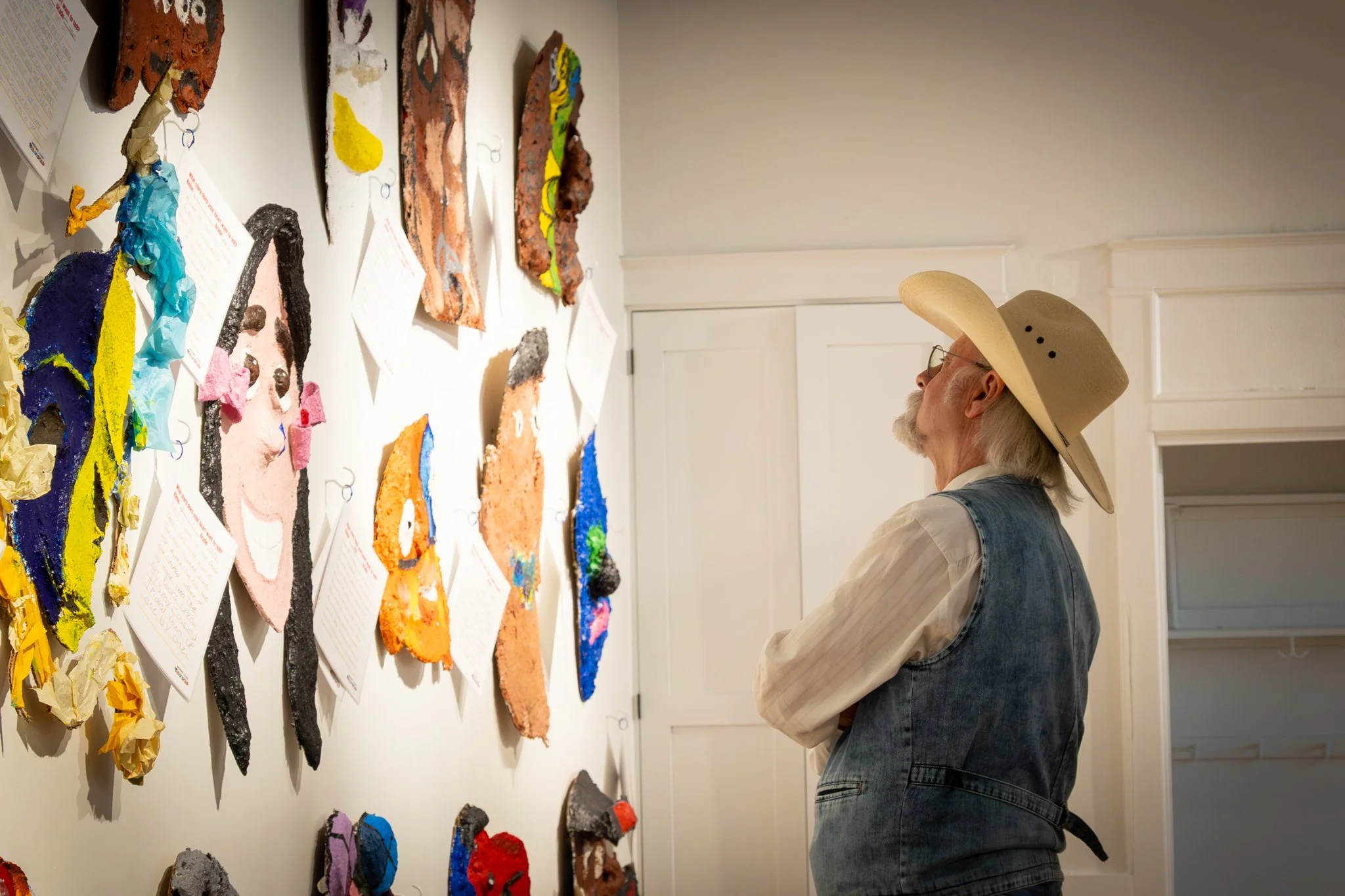 An older man with long white hair, glasses, and a beard, wearing a wide-brimmed hat inspects artwork on a gallery wall. The colorful art pieces are made from textured, torn paper and resemble abstract or stylized portraits, each accompanied by handwr