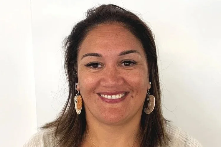 A woman with brown hair, smiling, wearing large earrings and a light-colored top, standing in front of a plain white background.