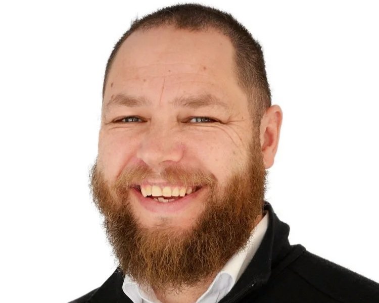 A smiling man with a beard and short hair, wearing a black jacket and white shirt, against a white background.