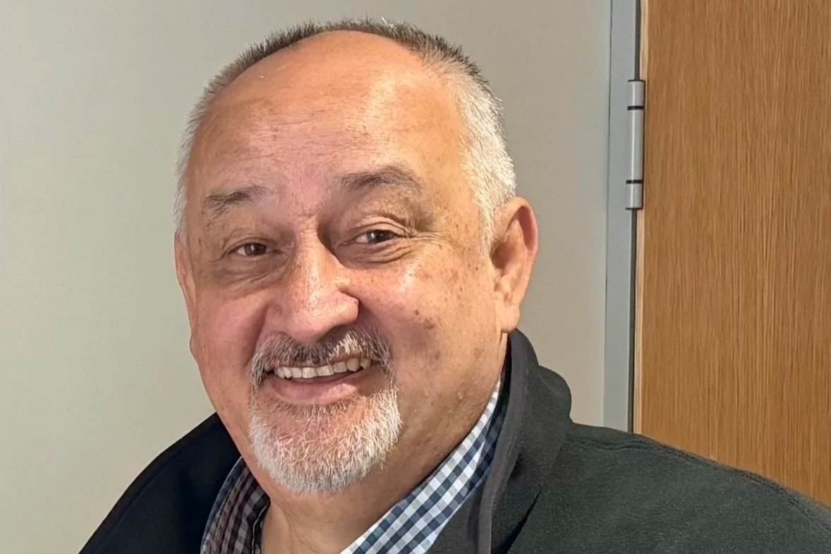 A smiling middle-aged man with short, gray hair, a beard, and a checked shirt, standing in front of a wooden door.