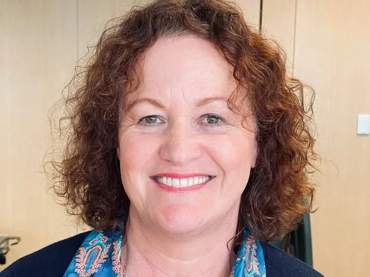 A smiling woman with curly reddish-brown hair, wearing a blue patterned scarf and dark top, in an indoor setting with a wooden wall behind her.