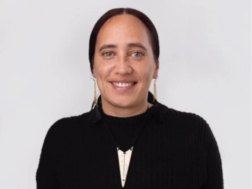 A woman with dark hair, light skin, and blue eyes smiling, wearing a black top, gold earrings, and a necklace with a large white pendant, standing against a plain white background.