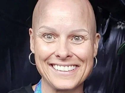 Close-up of a smiling woman with a shaved head wearing hoop earrings and a lanyard.