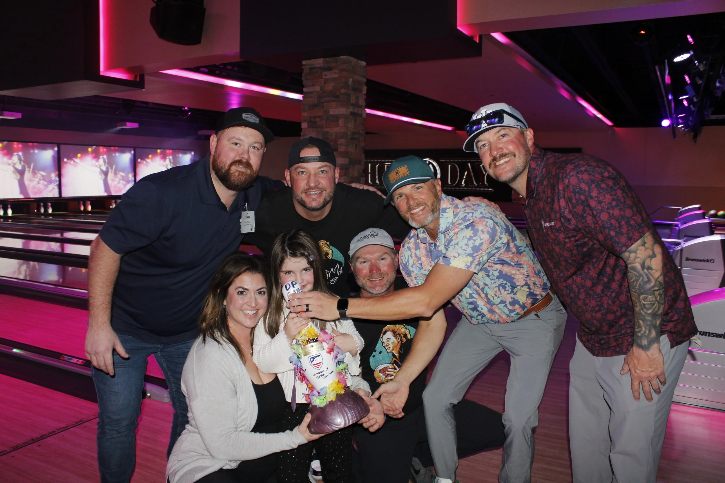 A group of seven people, including three women and four men, celebrating at a bowling alley. They are gathered around a young girl holding a decorated birthday cake, with smiles on their faces. The background shows bowling lanes and screens with colo