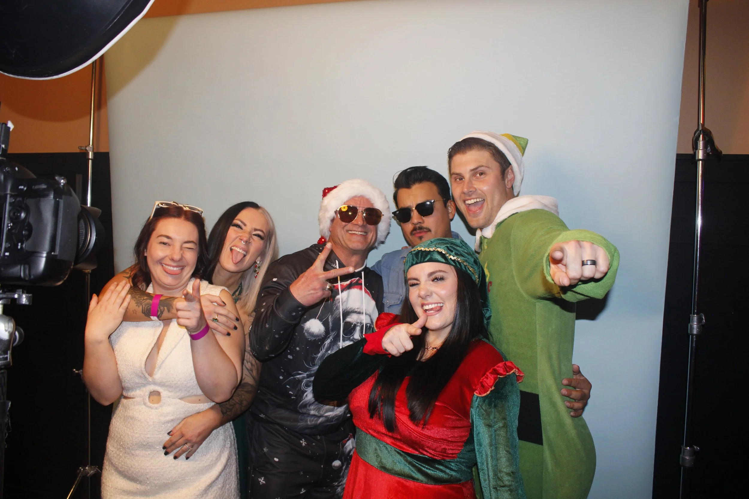 Group of six people dressed in festive holiday costumes and accessories, posing and smiling in front of a light blue background.