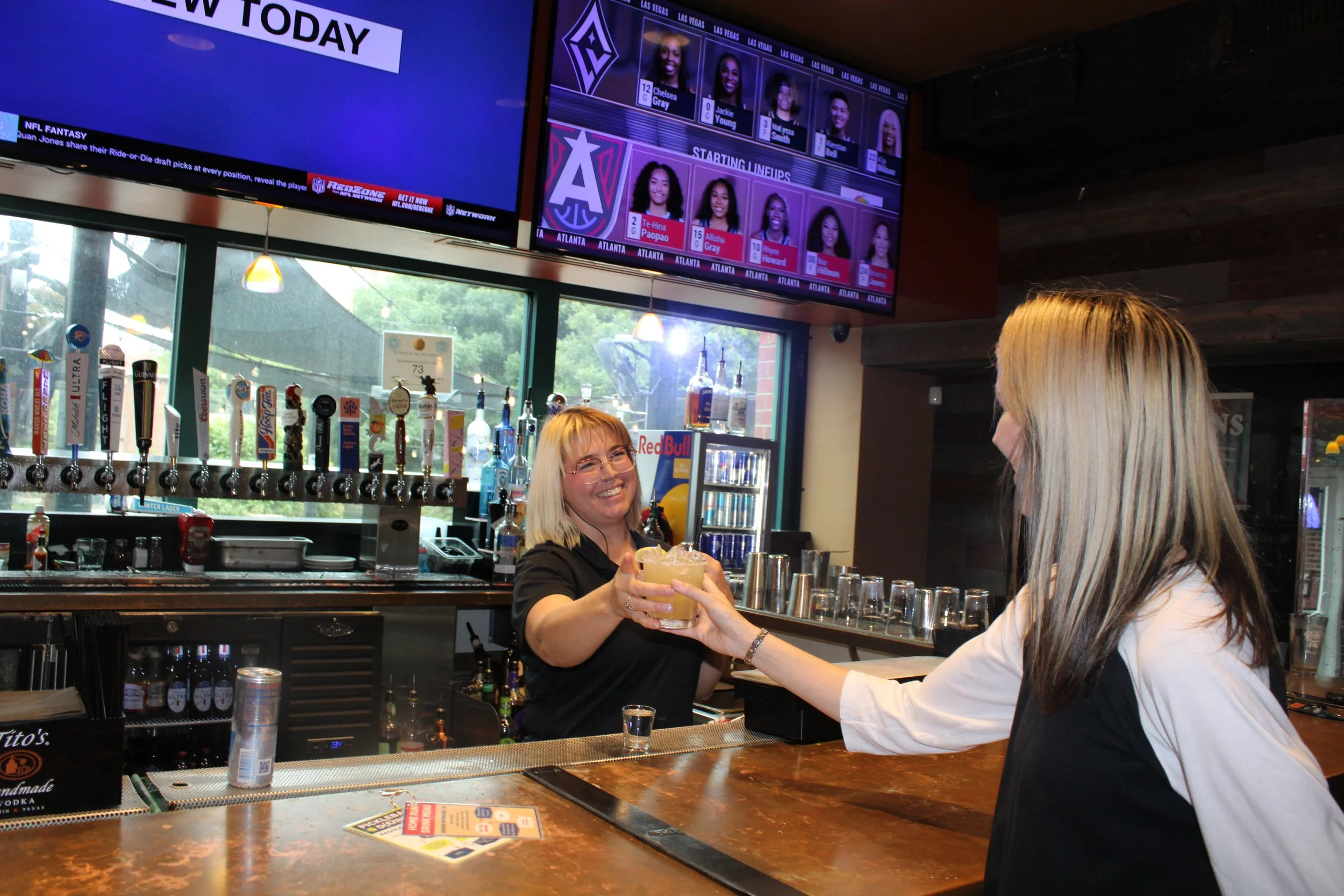 A bartender hands a drink to a smiling female customer in a bar with multiple taps and a large television screen displaying sports.
