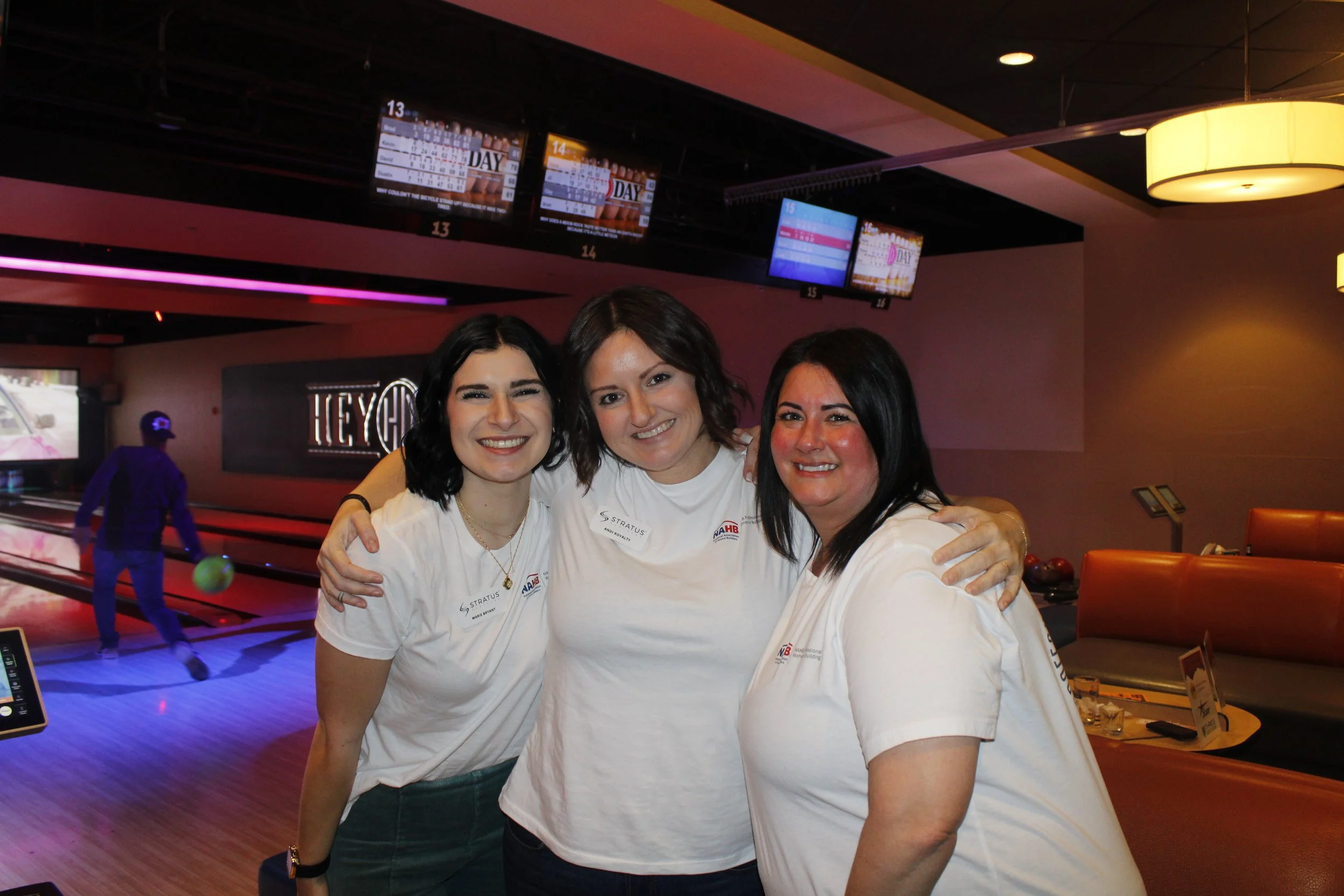 Three women smiling and hugging at a bowling alley, with a man bowling in the background and bowling lanes and digital scoreboards overhead.