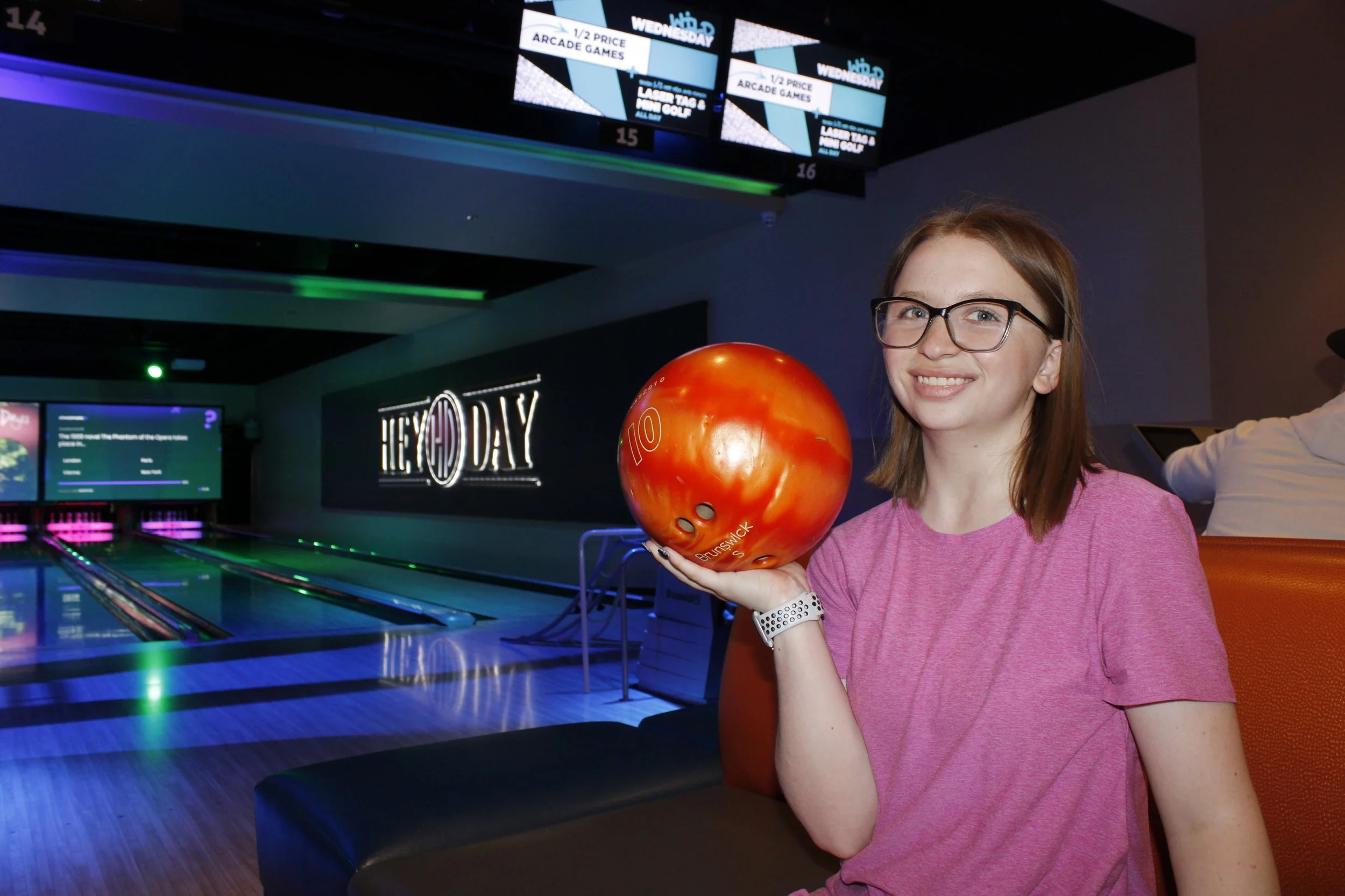 A young woman with glasses and a pink shirt smiling while holding a red bowling ball in a bowling alley.