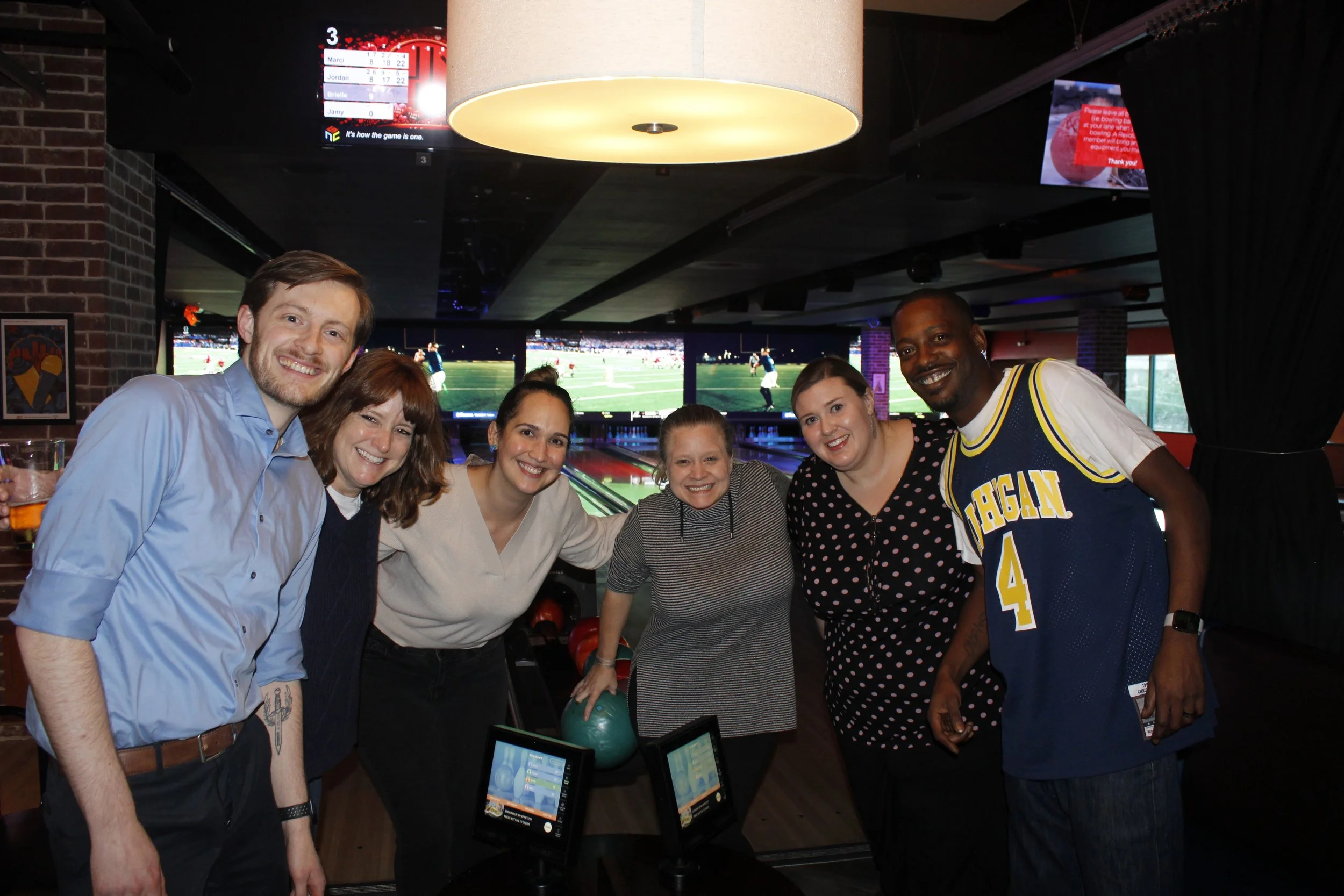 A group of six people smiling at a bowling alley, standing together near bowling balls and screens showing a sports game in the background.