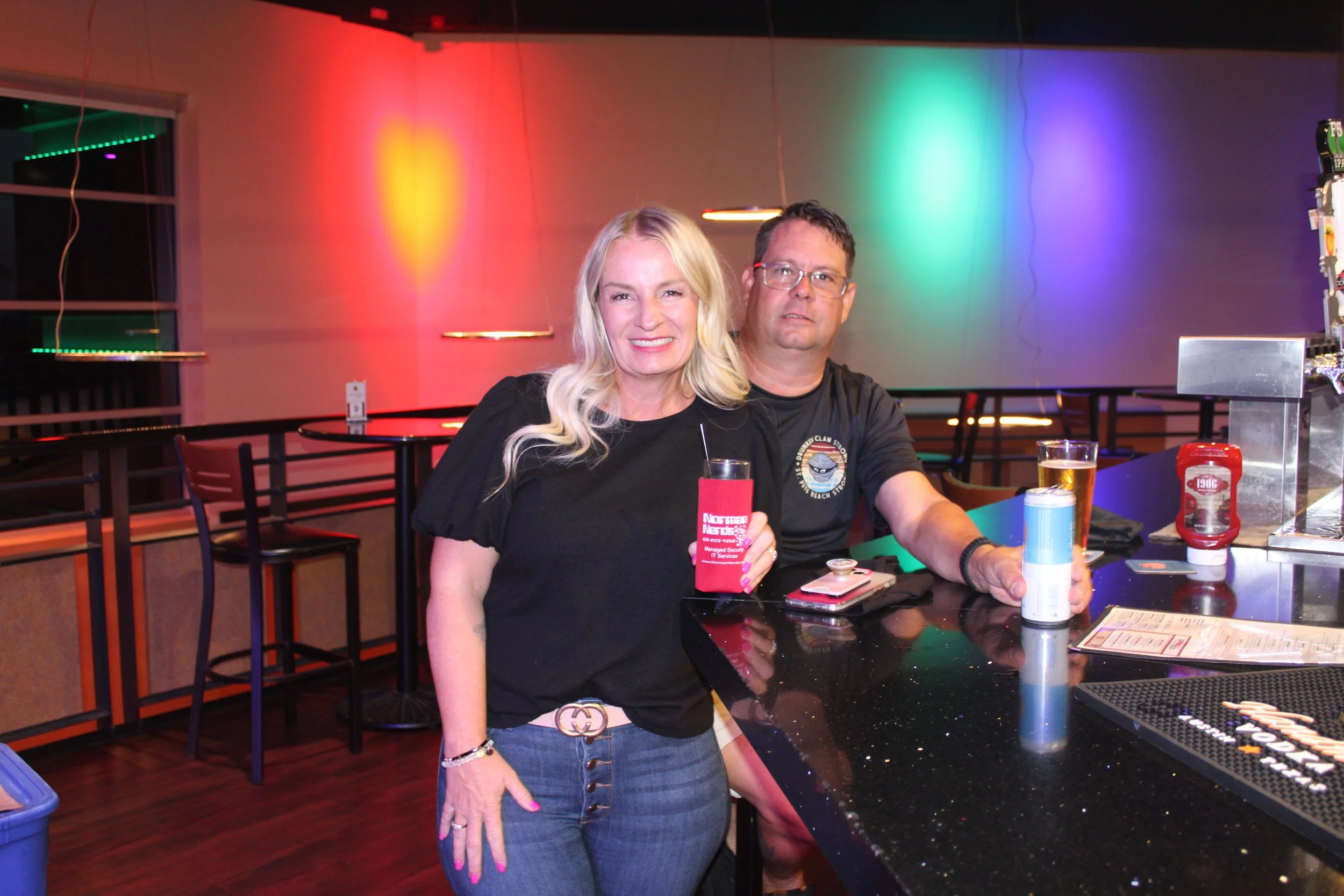 A woman and a man sitting at a bar counter with colorful neon lights in the background. The woman is smiling, holding a drink, wearing a black top, blue jeans, and a Gucci belt. The man is seated next to her with a beer in hand, wearing glasses and a black T-shirt. The bar counter has condiments, menus, and drink coasters.