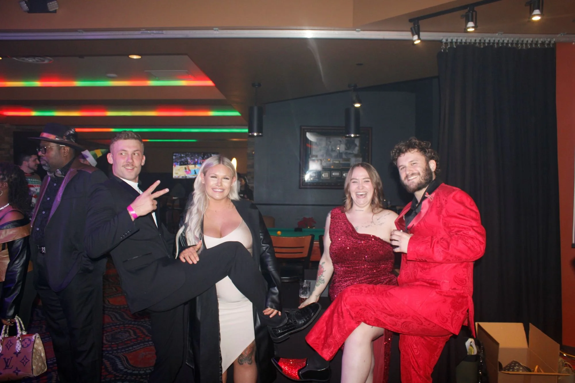 Group of five people at a party, standing in front of a pool table, smiling and posing for the camera; colorful lighting and a TV screen in the background.