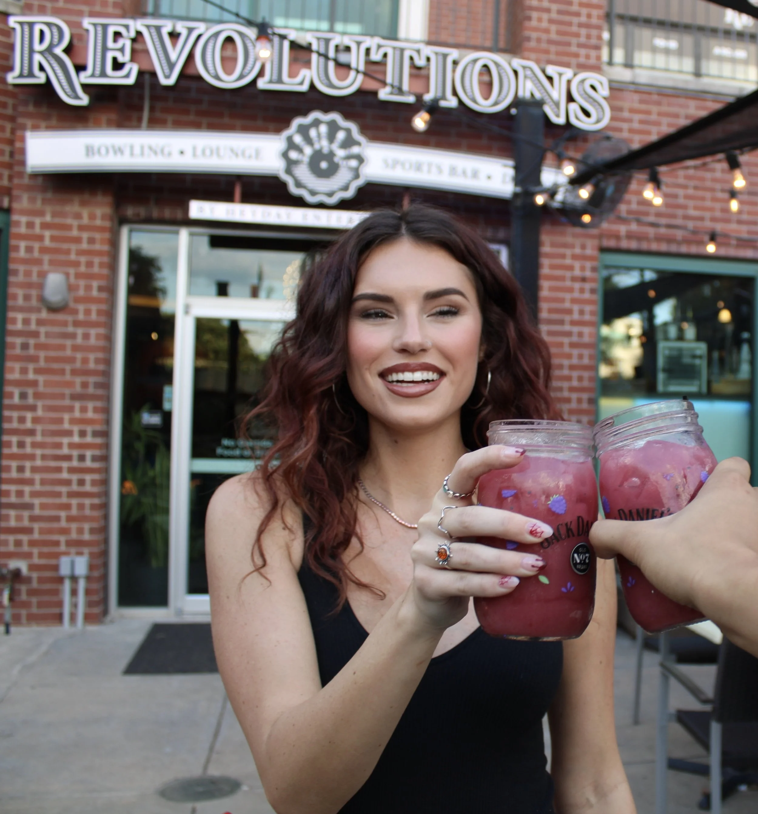 A woman with curly red hair smiling and holding a jar of pink drink, standing outside a brick building with a sign reading 'Revolutions' and 'Bowling Lounge'.