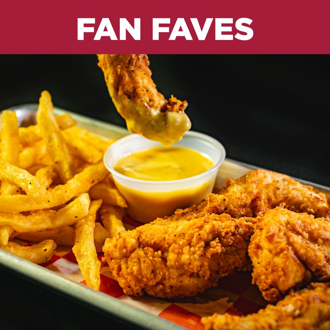 A plate of fried chicken tenders, French fries with seasoning, a small container of dipping sauce, and a tall glass of soda on a checkered paper at a bar or restaurant.