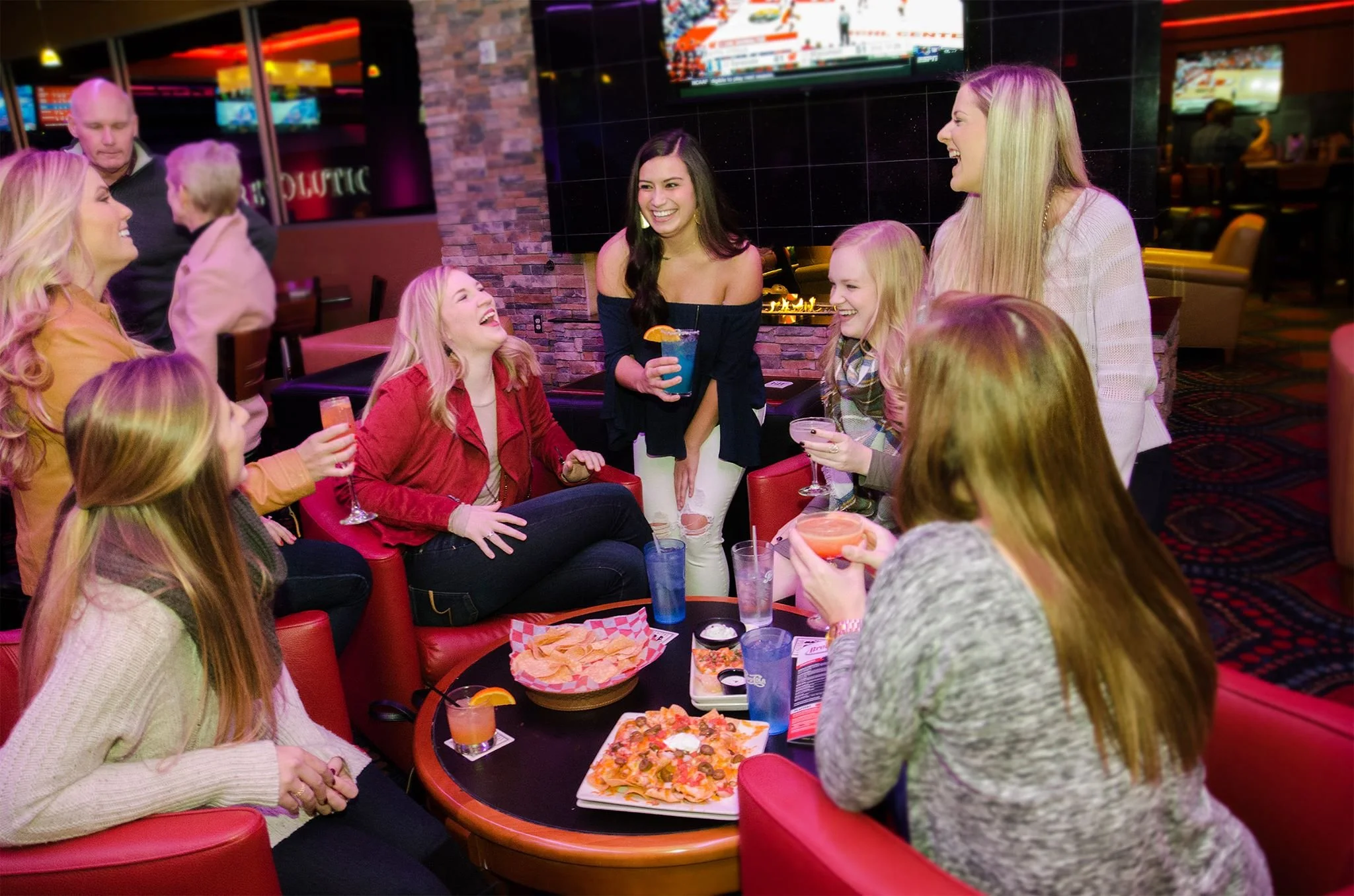 A group of women socializing and laughing at a restaurant or bar, with food and drinks on the table and a large screen TV in the background.