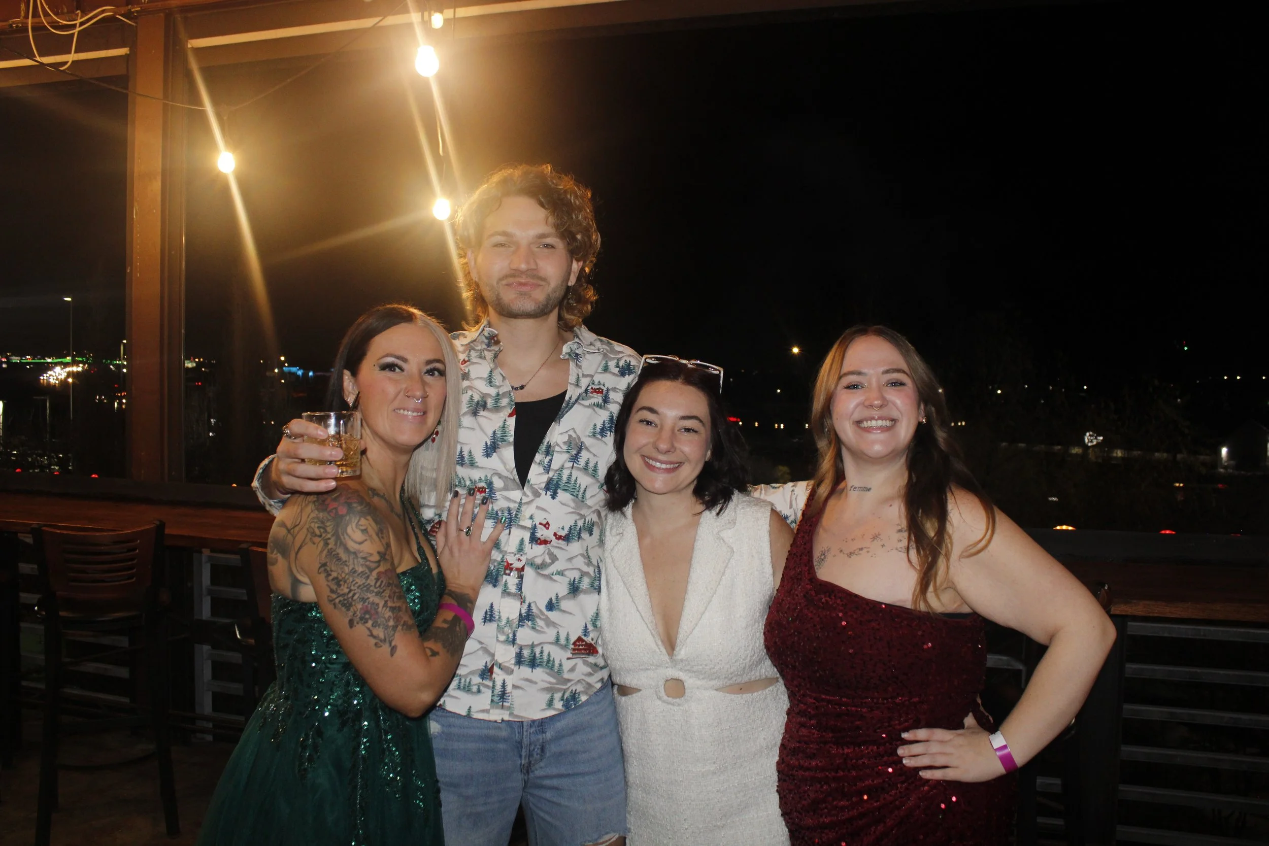 Group of four friends smiling and posing at night on a balcony, with city lights in the background. One woman has tattoos and holds a drink, the man and women wear formal attire.