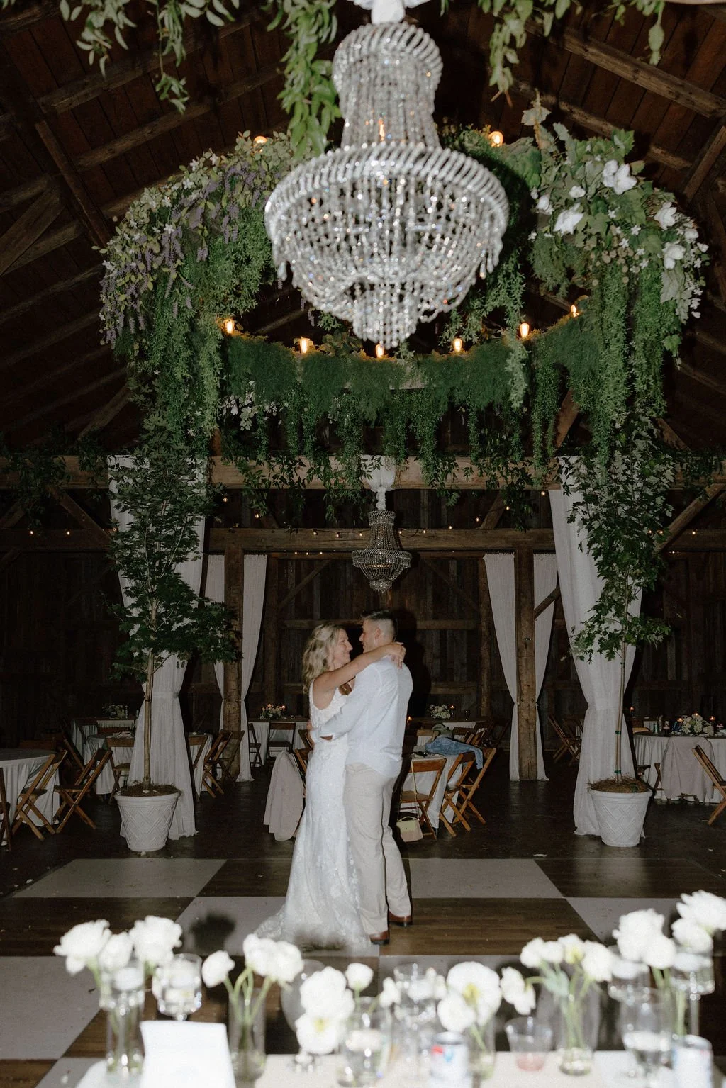 Image of a bride and groom having a private dance.  The room features a checkered dancefloor and heavy greenery, including potted maple trees and installed vines.