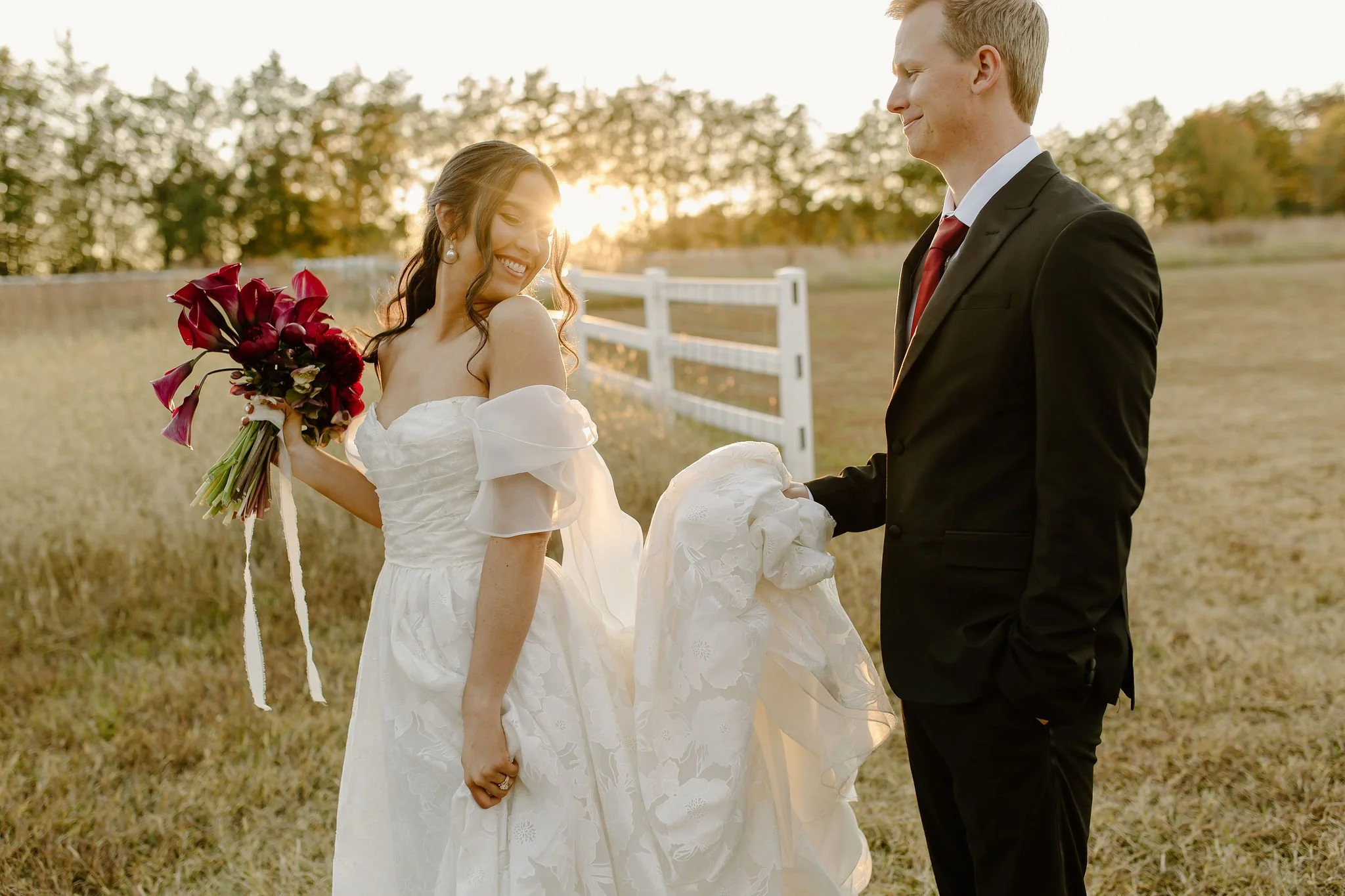 A bride and groom on a farm at sunset, with the bride holding a bouquet of red and purple flowers and the groom holding a piece of her wedding dress.