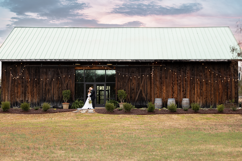 A bride and groom standing in front of a rustic wooden barn, decorated with string lights and surrounded by small bushes, during sunset.