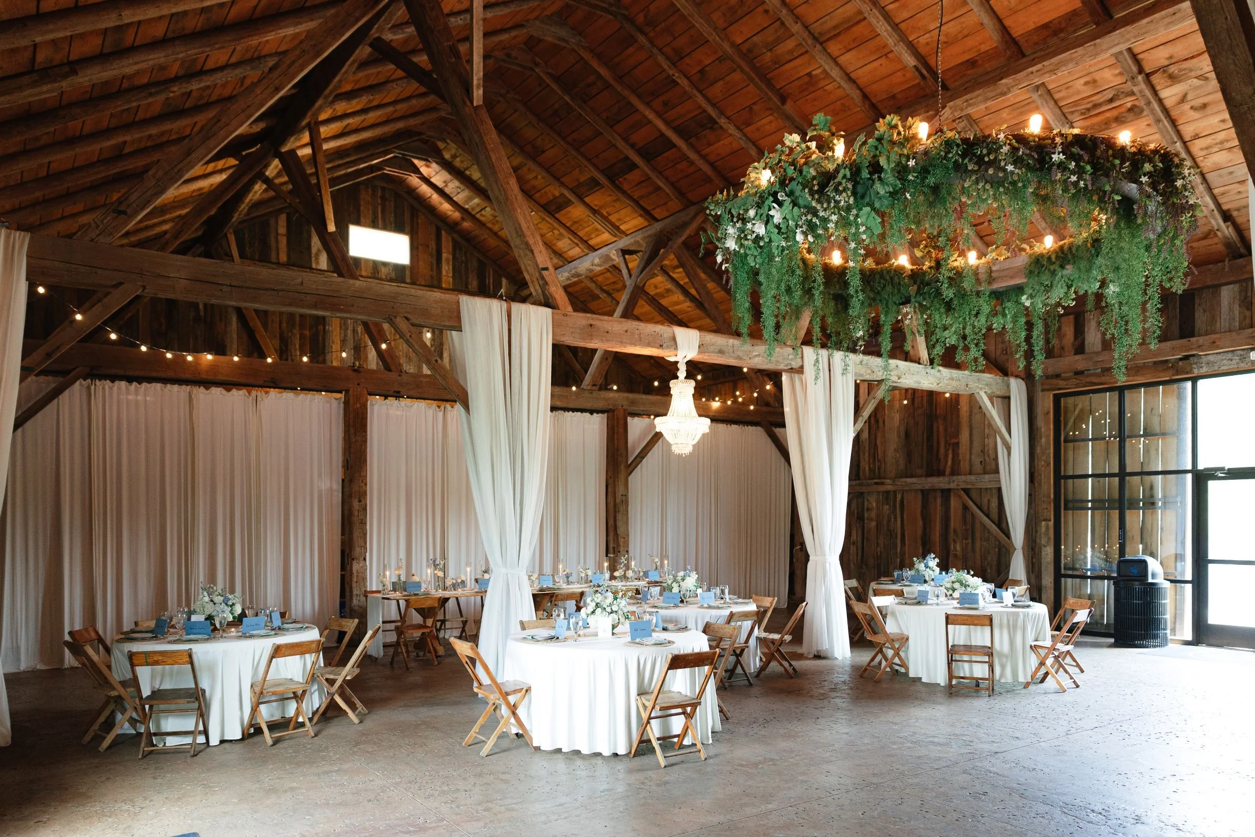 Interior of a rustic barn decorated for a wedding reception with round white tablecloths, floral centerpieces, and string lights, featuring wooden beams and a hanging floral chandelier.