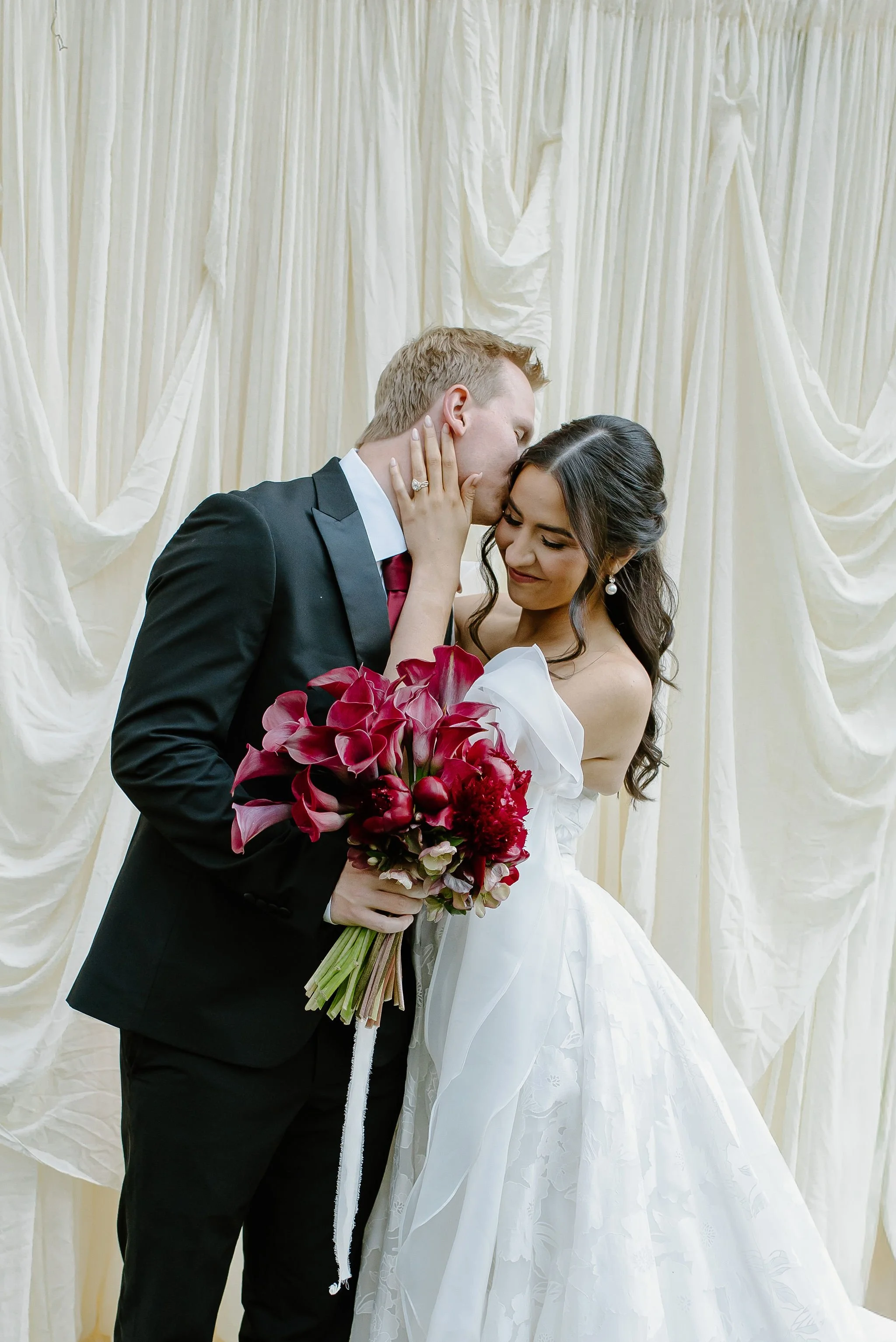 A newlywed couple sharing a kiss on their wedding day, with the groom gently holding the bride and the bride holding a bouquet of red and purple flowers.