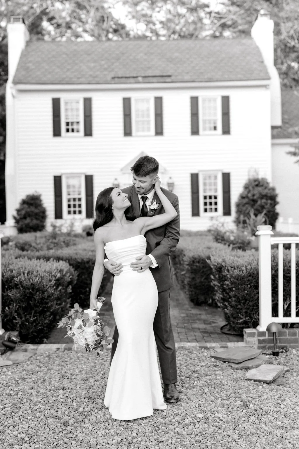 A newlywed couple standing outside a house, smiling at each other, with the bride holding a bouquet and the groom dressed in a suit.
