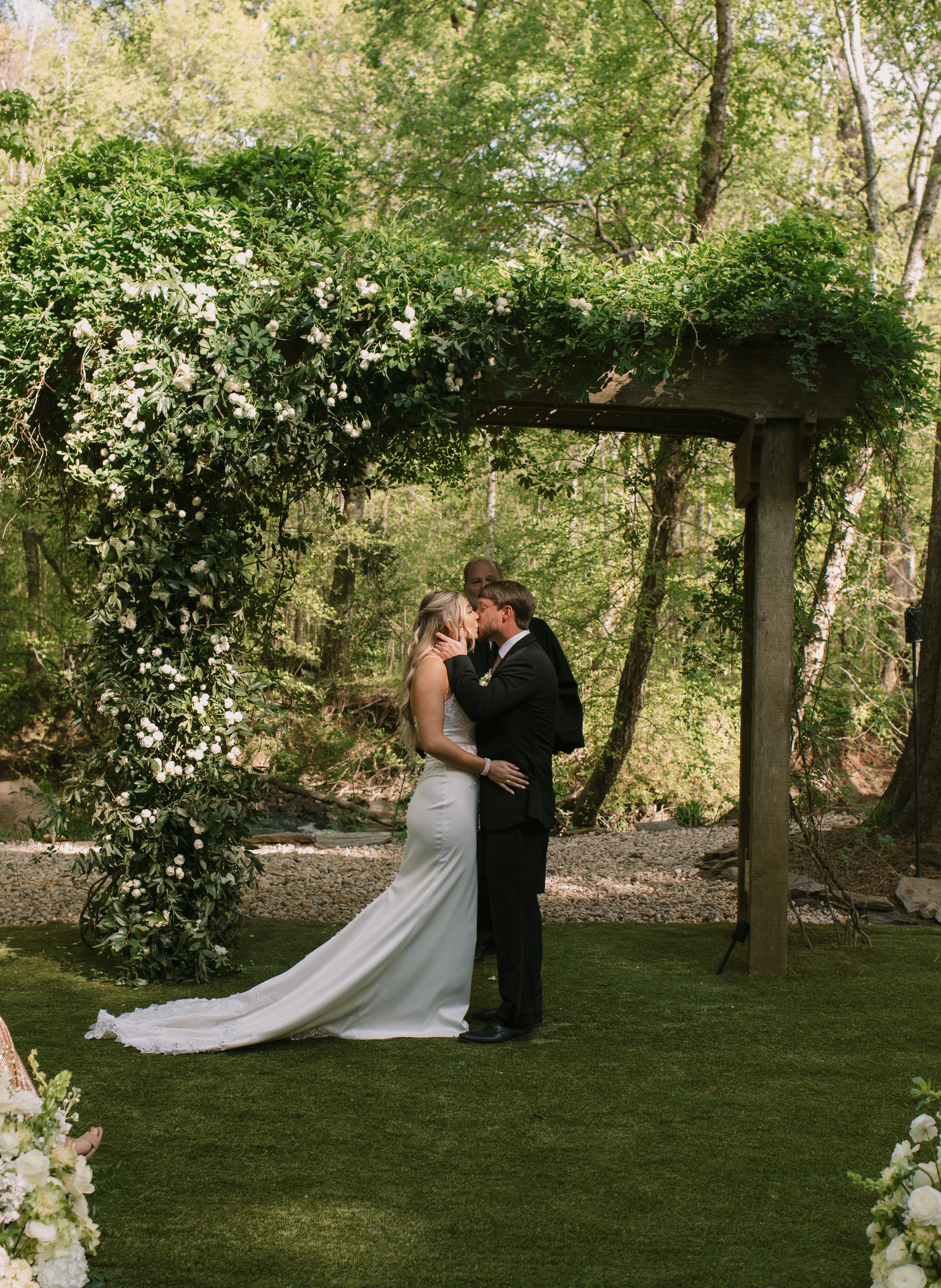 A bride and groom share a kiss during their wedding ceremony outdoors under a wooden arbor decorated with green leaves and white flowers, surrounded by trees and greenery.