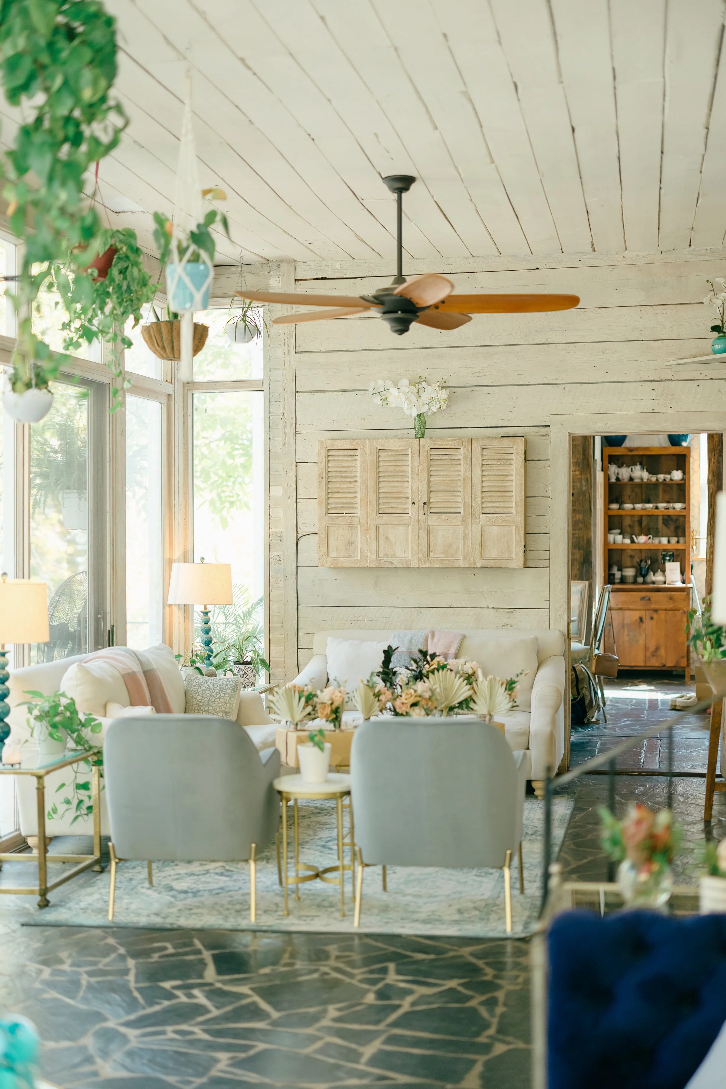 Bright indoor living room with white wooden walls and ceiling, large windows, beige sofas with pillows, a glass coffee table with flowers, hanging plants, a ceiling fan, and a wooden cabinet.
