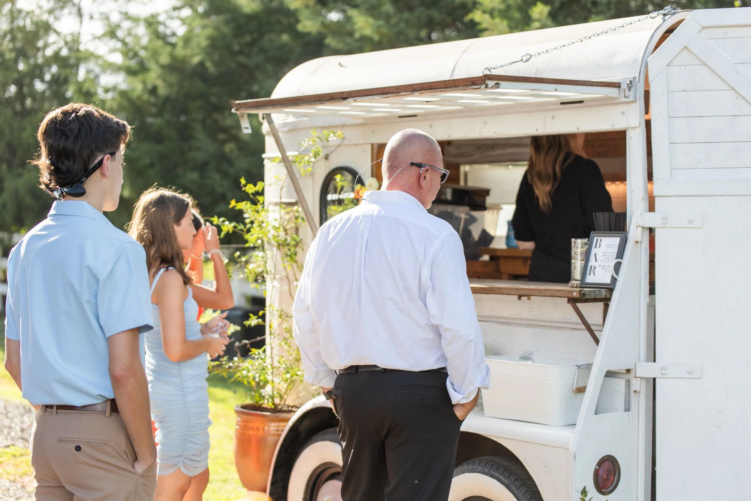 People standing in line at a white food truck serving drinks outdoors on a sunny day.