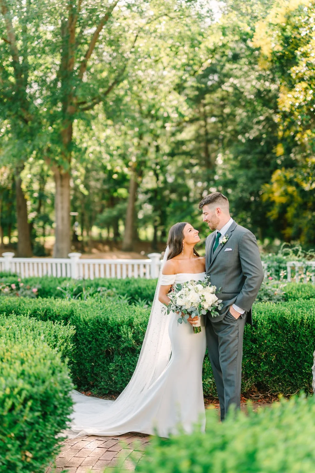 A bride and groom stand outdoors in a lush, green garden with trees and bushes, looking into each other's eyes. The bride wears a white off-shoulder wedding gown and holds a bouquet of white flowers, while the groom wears a gray suit with a tie and a