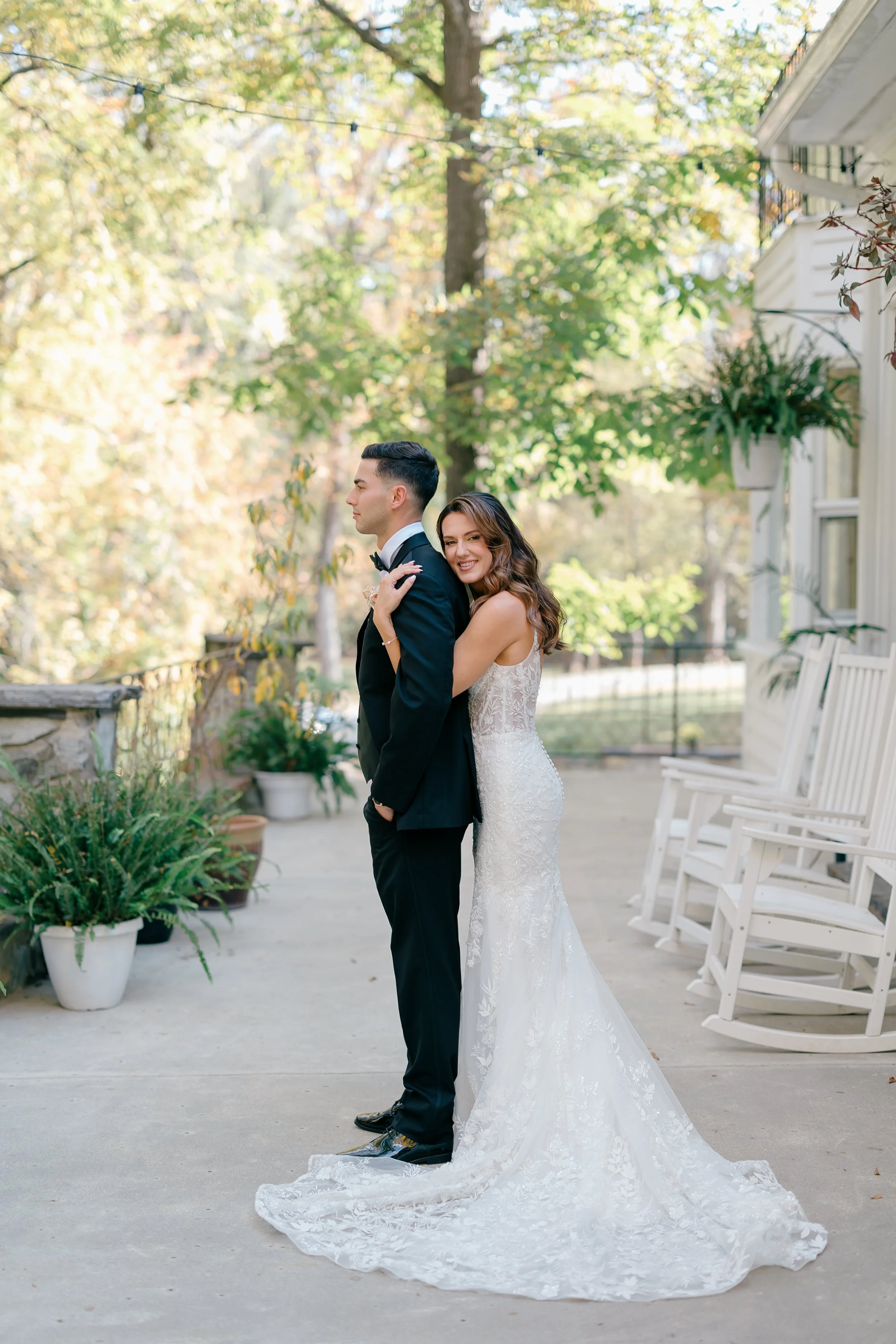 Bride and groom standing outdoors on a wedding day. The bride is smiling and hugging the groom from behind, who is facing sideways. She wears a white lace wedding dress with a long train, and he wears a black tuxedo with a bow tie. The scene is set i