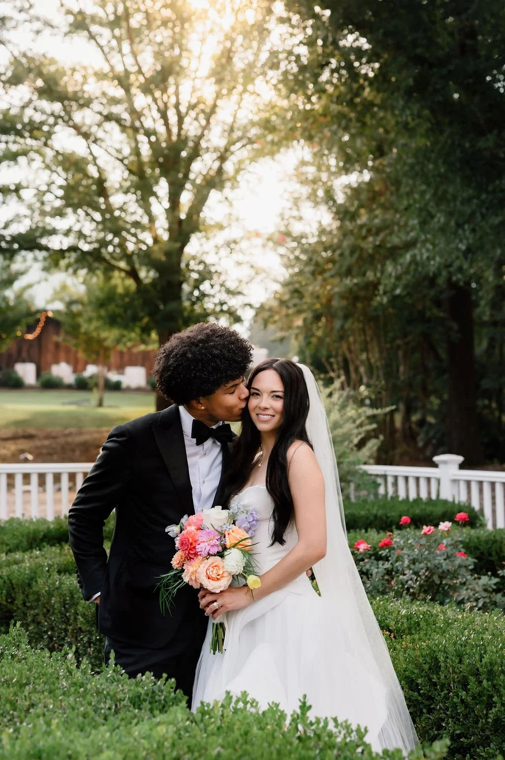A newlywed couple standing in a garden, with the groom kissing the bride on the cheek. The bride is holding a colorful bouquet of flowers, and they are smiling. The setting is outdoors with greenery and sunlit trees in the background.