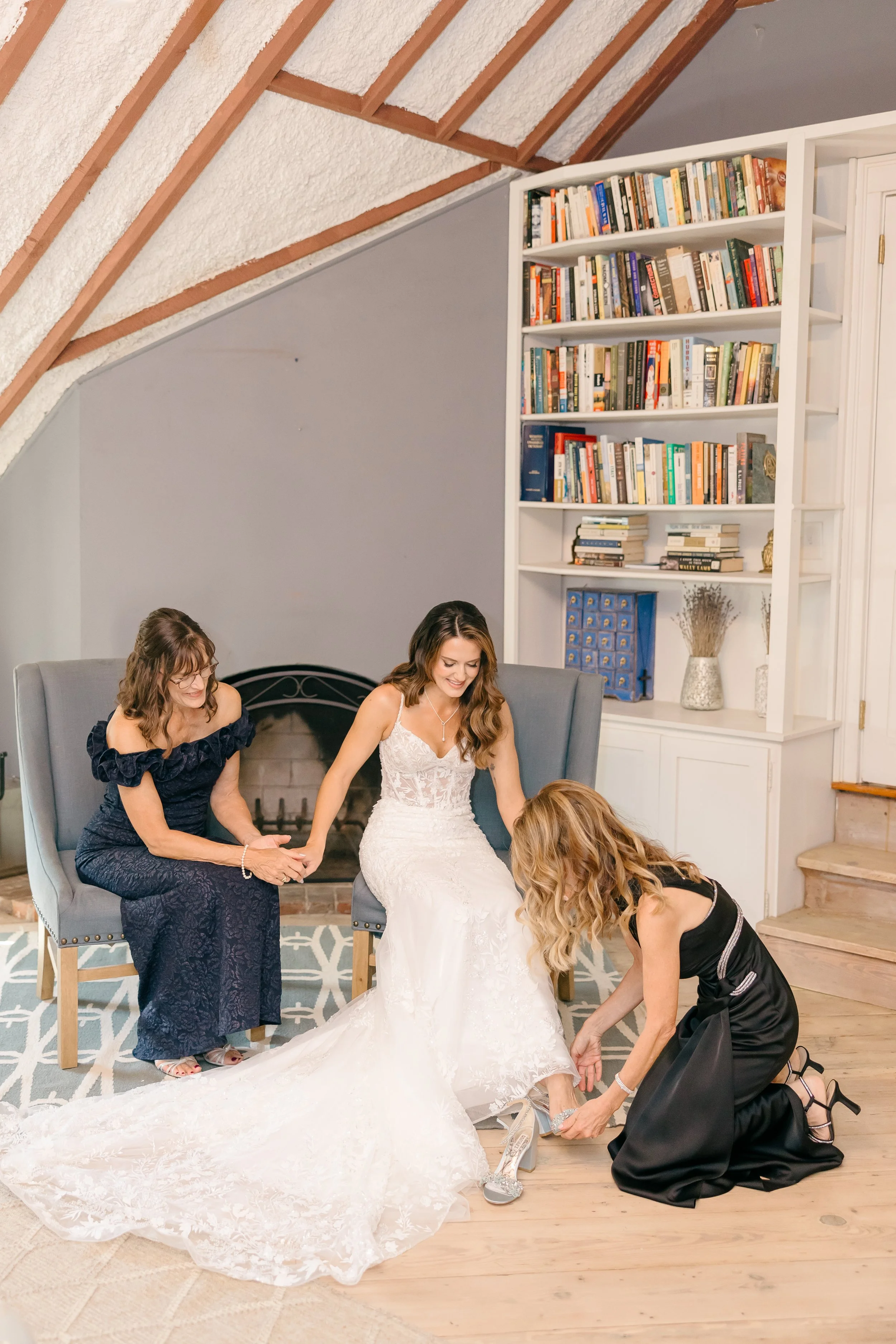 A bride in a white wedding dress sitting on a chair while friends help her put on her shoes in a cozy living room with a gray and white color scheme and a large bookshelf in the background.