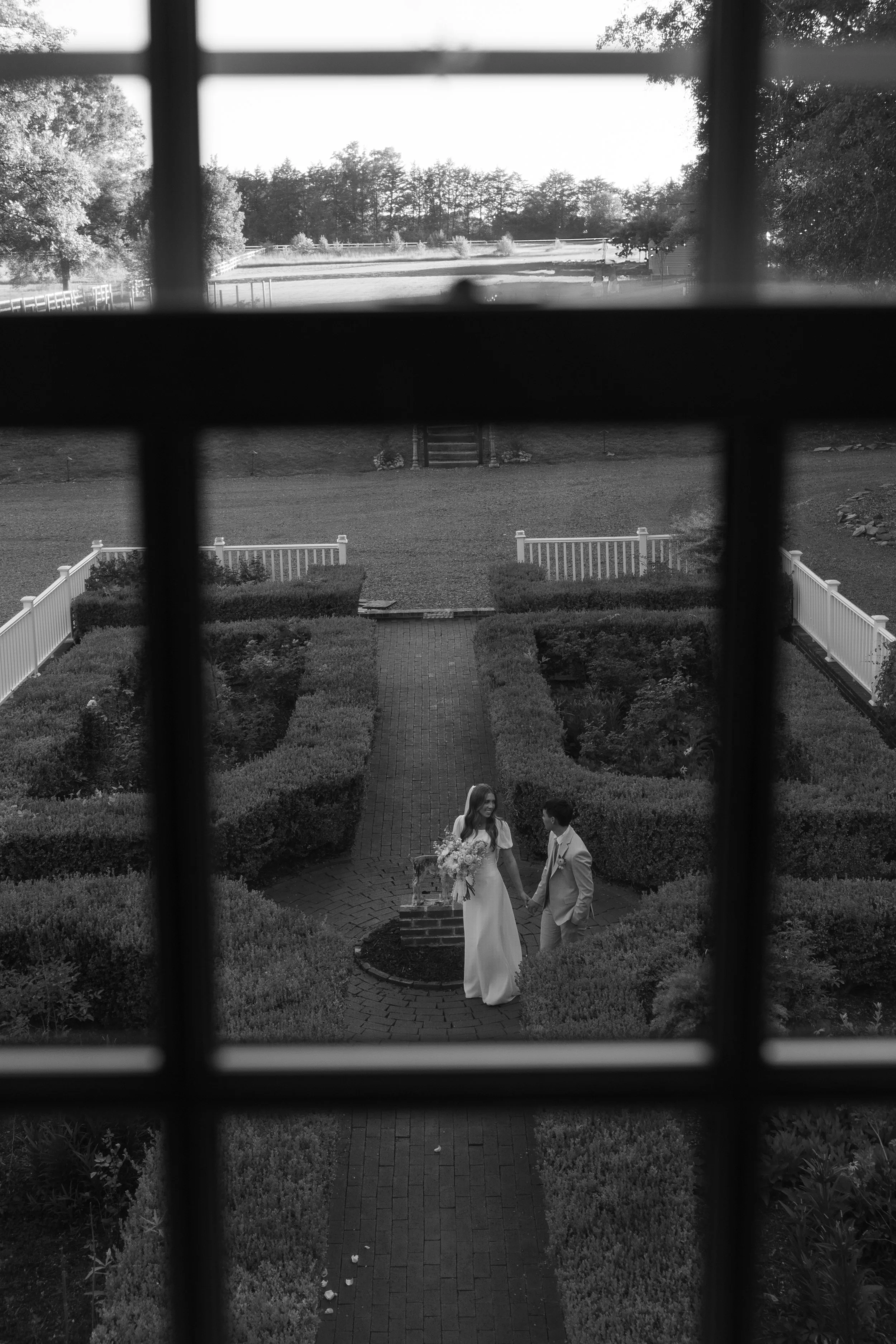 A bride and groom holding hands and smiling at each other outside in a garden, viewed through a window with grid pattern, with well-manicured bushes and a brick pathway.