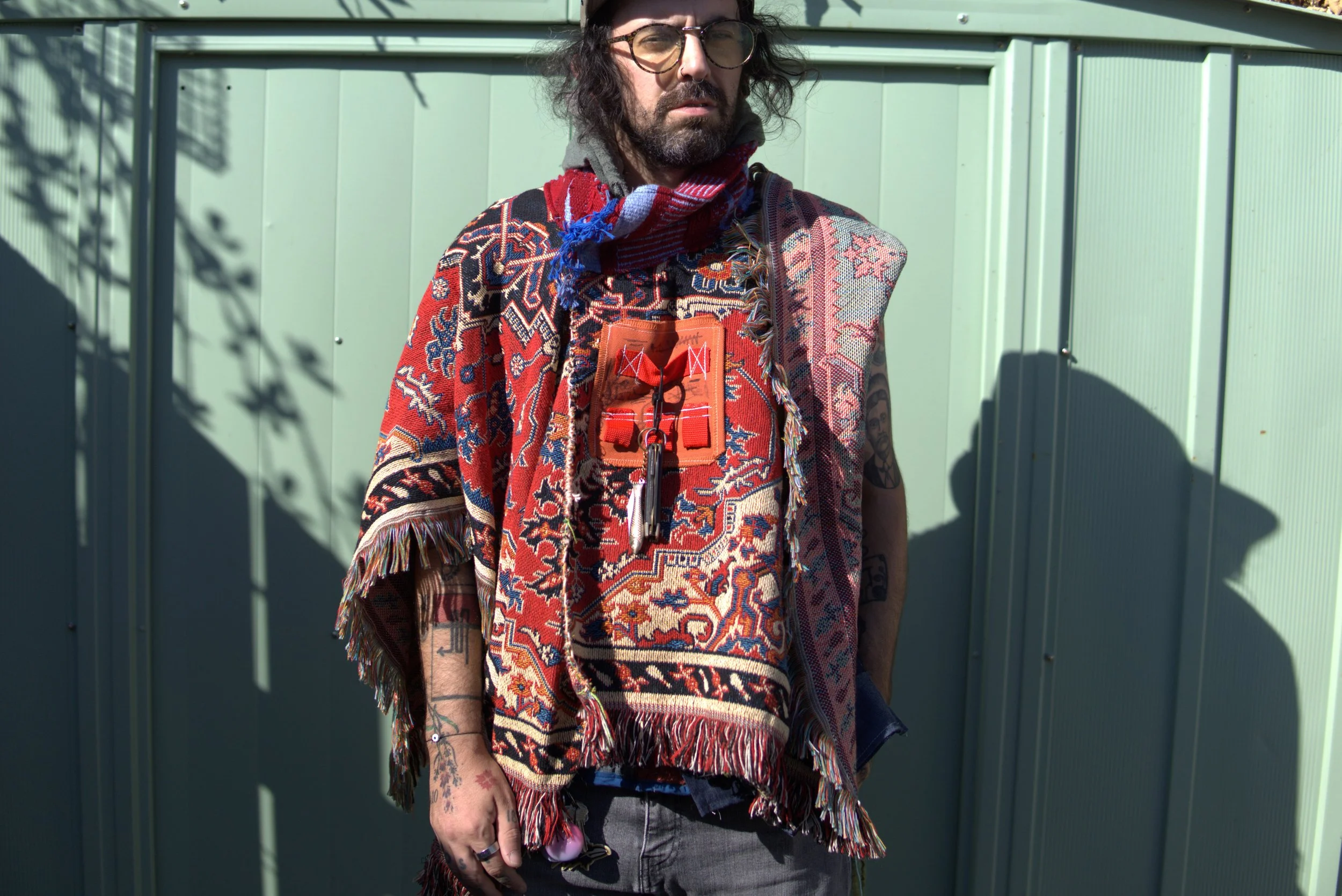 A man with long dark hair, glasses, and tattoos wearing a colorful patterned poncho, a red and blue scarf, and a necklace with keys, standing outdoors against a green wall with shadows of tree branches.