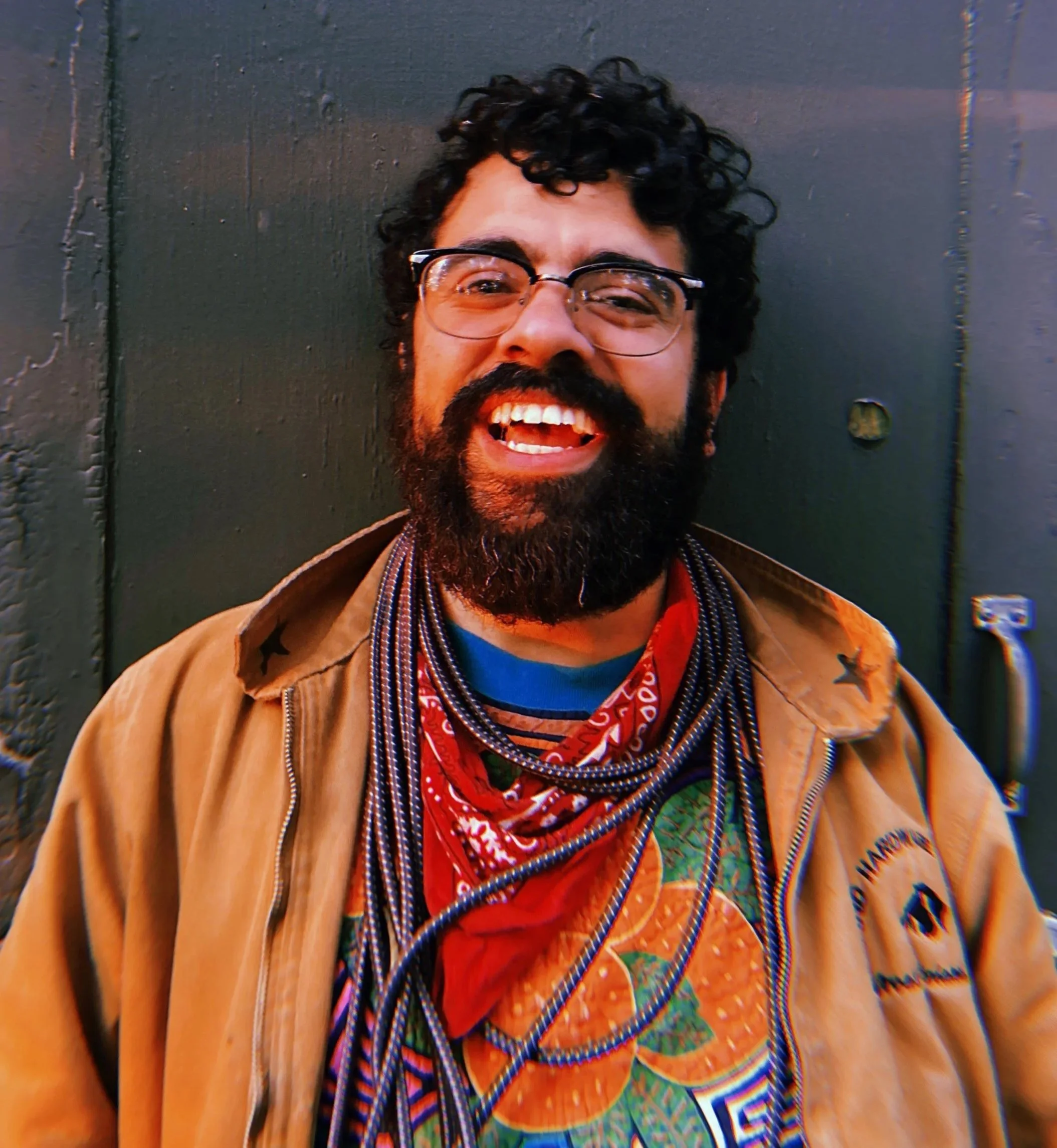 A man with glasses and a beard, smiling and showing teeth, standing against a black wall, wearing a tan jacket, colorful shirt, and red bandana.