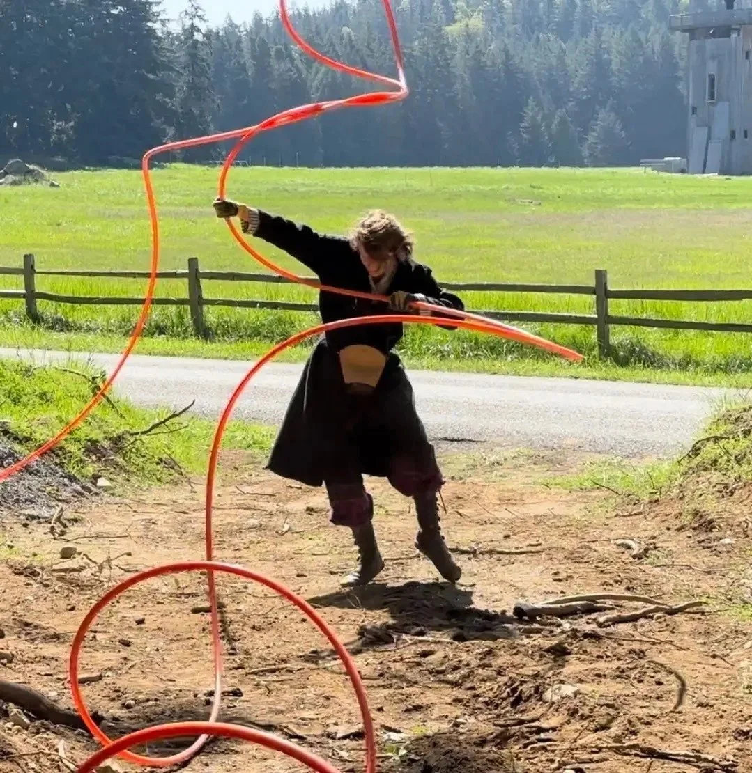 A person with long hair installing electrical conduit while dancing, in a grassy and dirt area with trees and a building in the background.