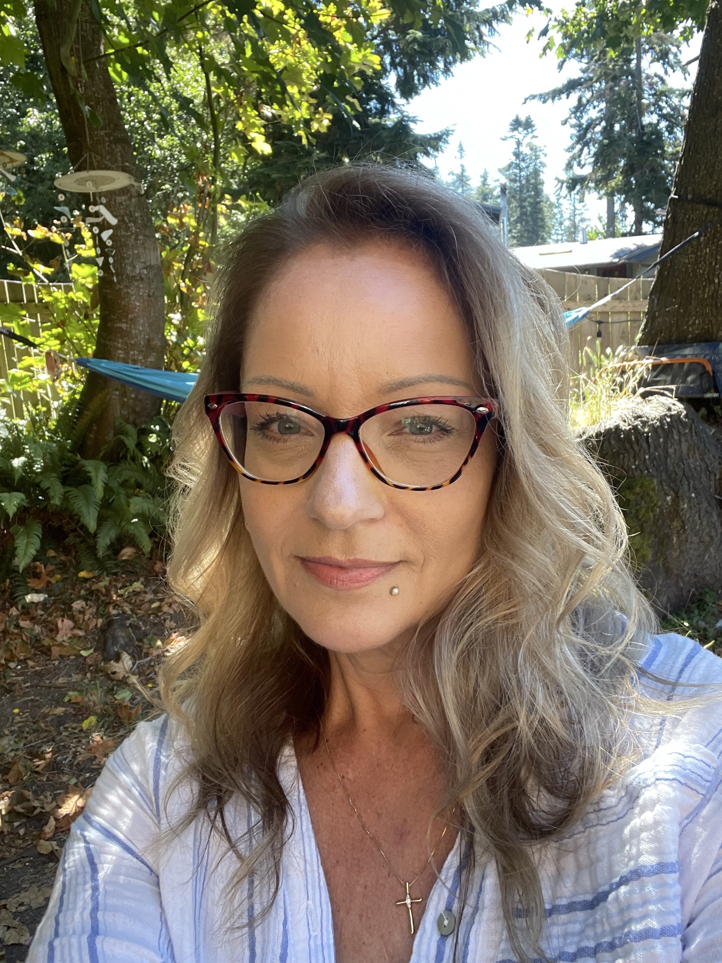 A woman with glasses and a small piercing on her lower lip takes a selfie outdoors in a backyard with trees, plants, and a hammock in the background.