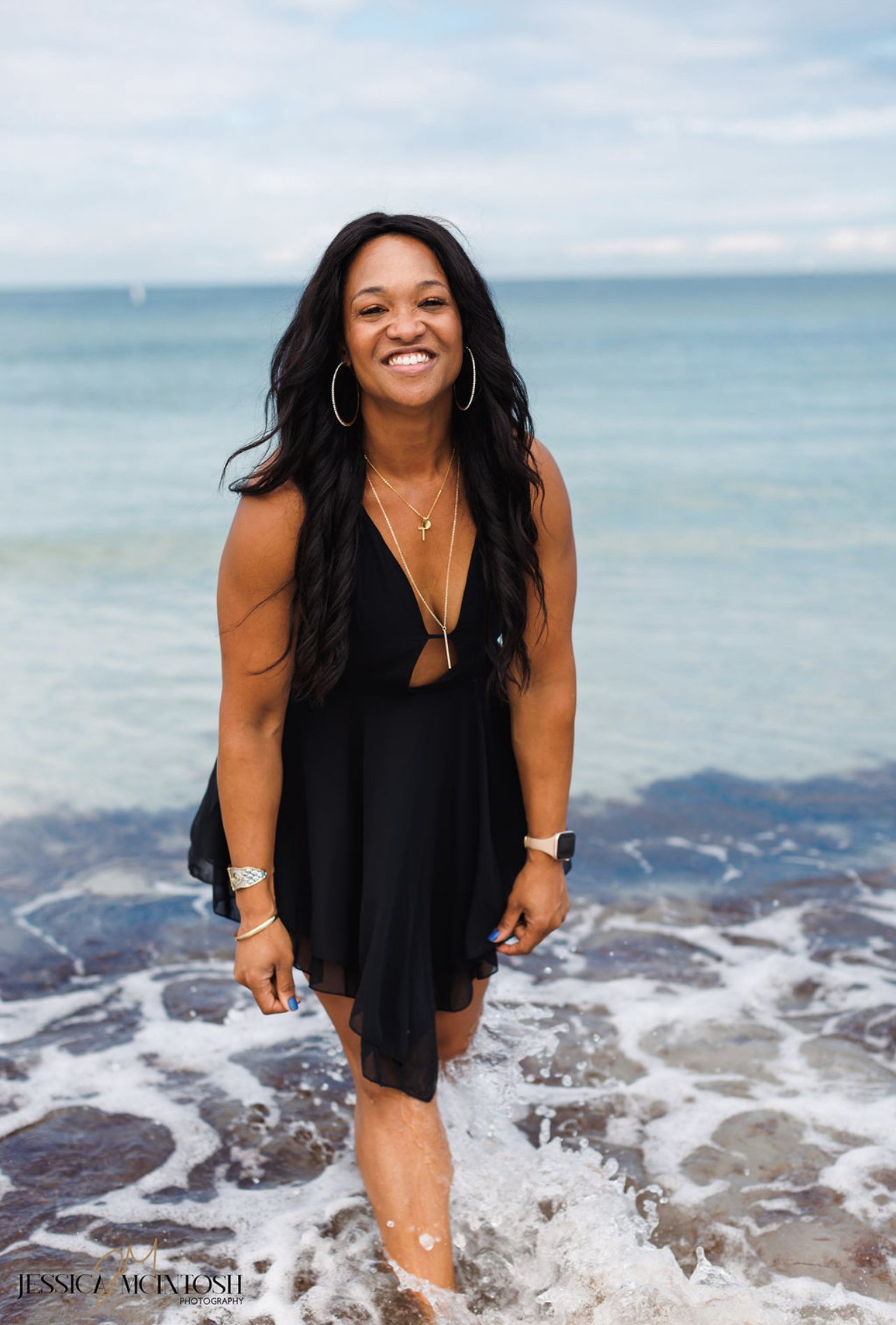 Woman in black dress smiling by the sea, with waves touching her feet.