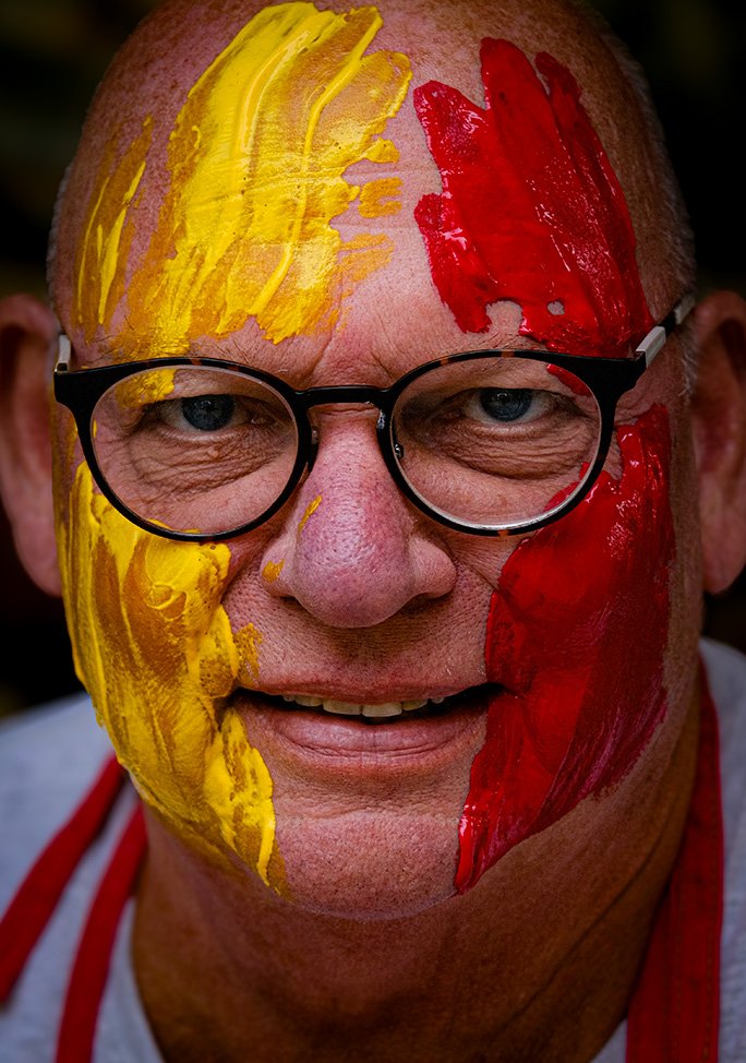 A man with painted yellow and red stripes on his face is wearing glasses and smiling.