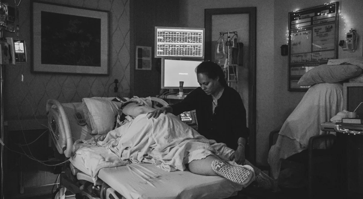 Black and white photo of a laboring mom on her side with an epidural, in side lying release while doula Jennifer Bianco of A Joyful Blessing Birth Services assists her in achieving an optimal position for baby during a hospital birth.