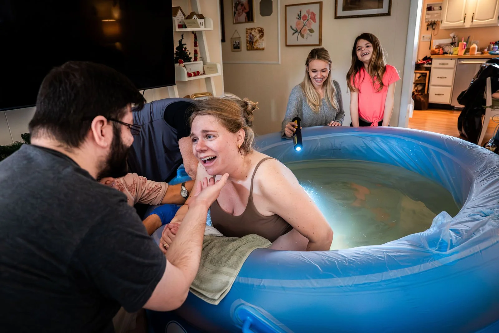 Mom smiling in a birthing tub as her husband gently cradles her face, with birth attendants-including midwives, her sister, and daughter-smiling in the background during a home birth.
